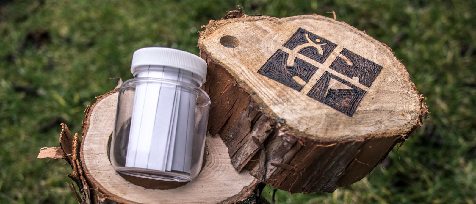 En tjock gren med ett gångjärn som gör att den går att öppna för att komma åt en burk som ligger i ett borrat hål i. På änden är det inbränt en geocachingsymbol