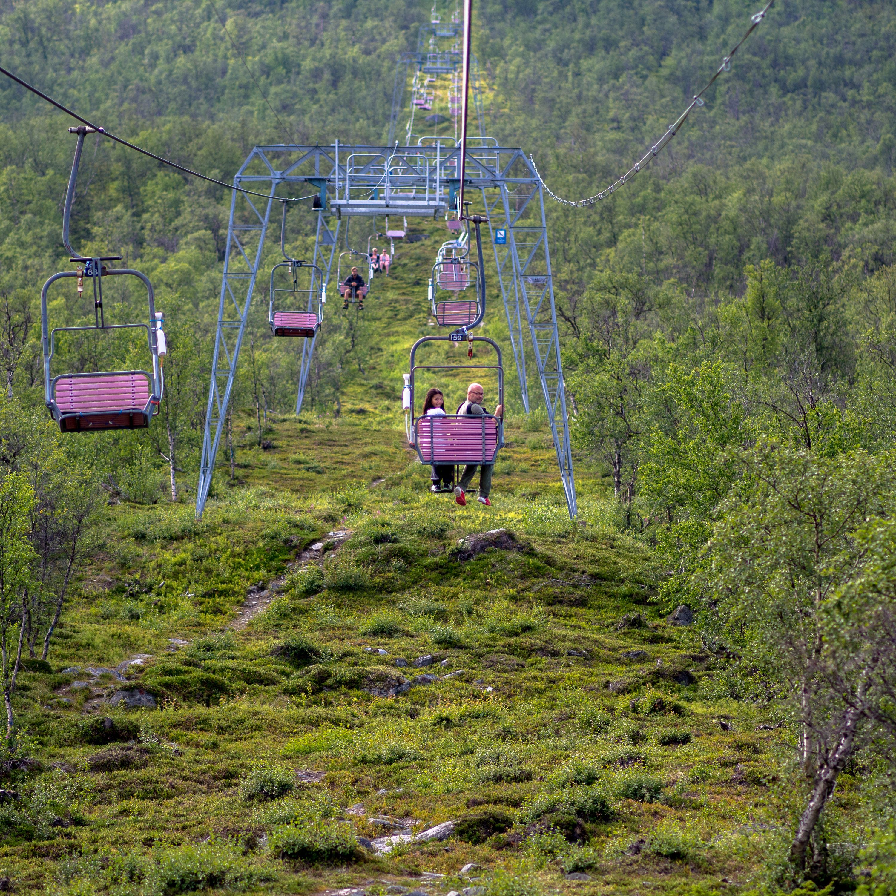 En linbana som går upp mot ett fjäll. Mitt i bild är det en stol med två personer som vänt på sig och tittar rakt in i kameran