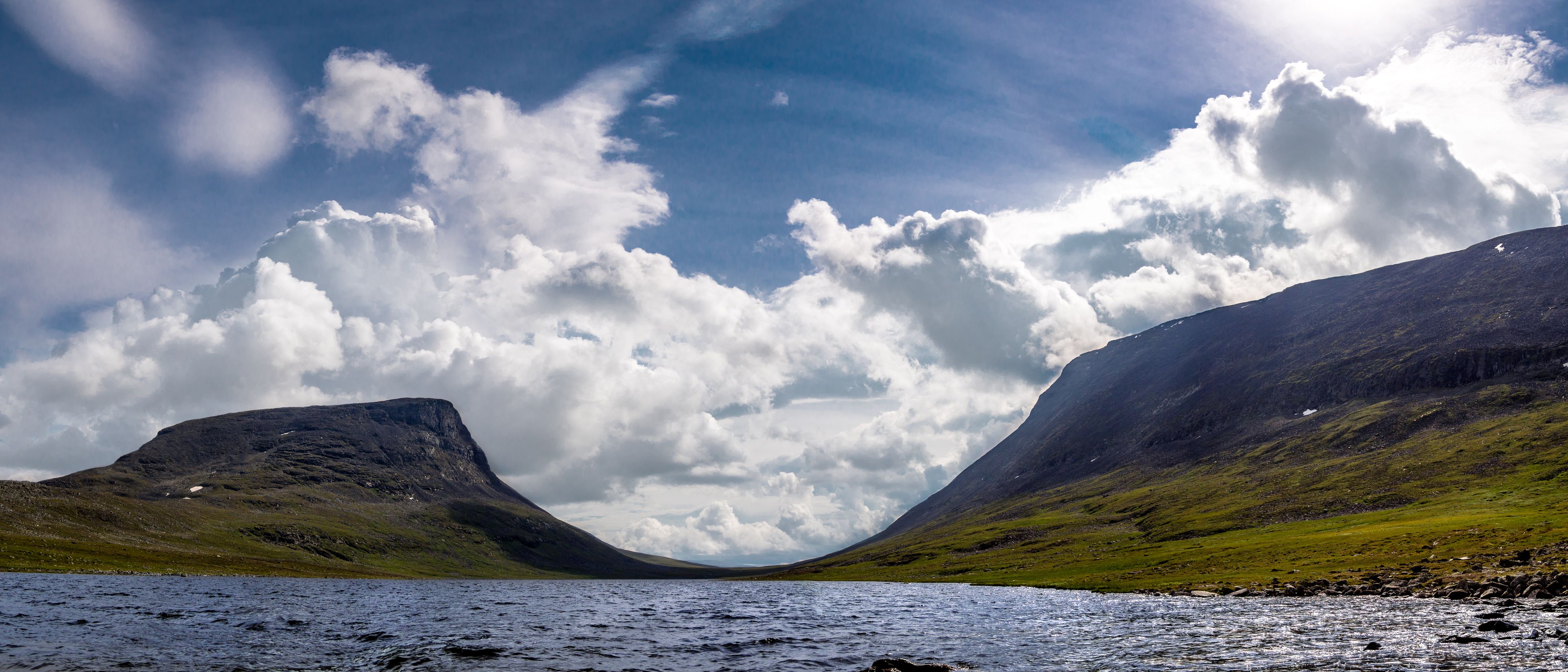 Lapporten under en blå himmel med vita moln längs horisonten. Framför Lapporten syns sjön Čuonjájávri