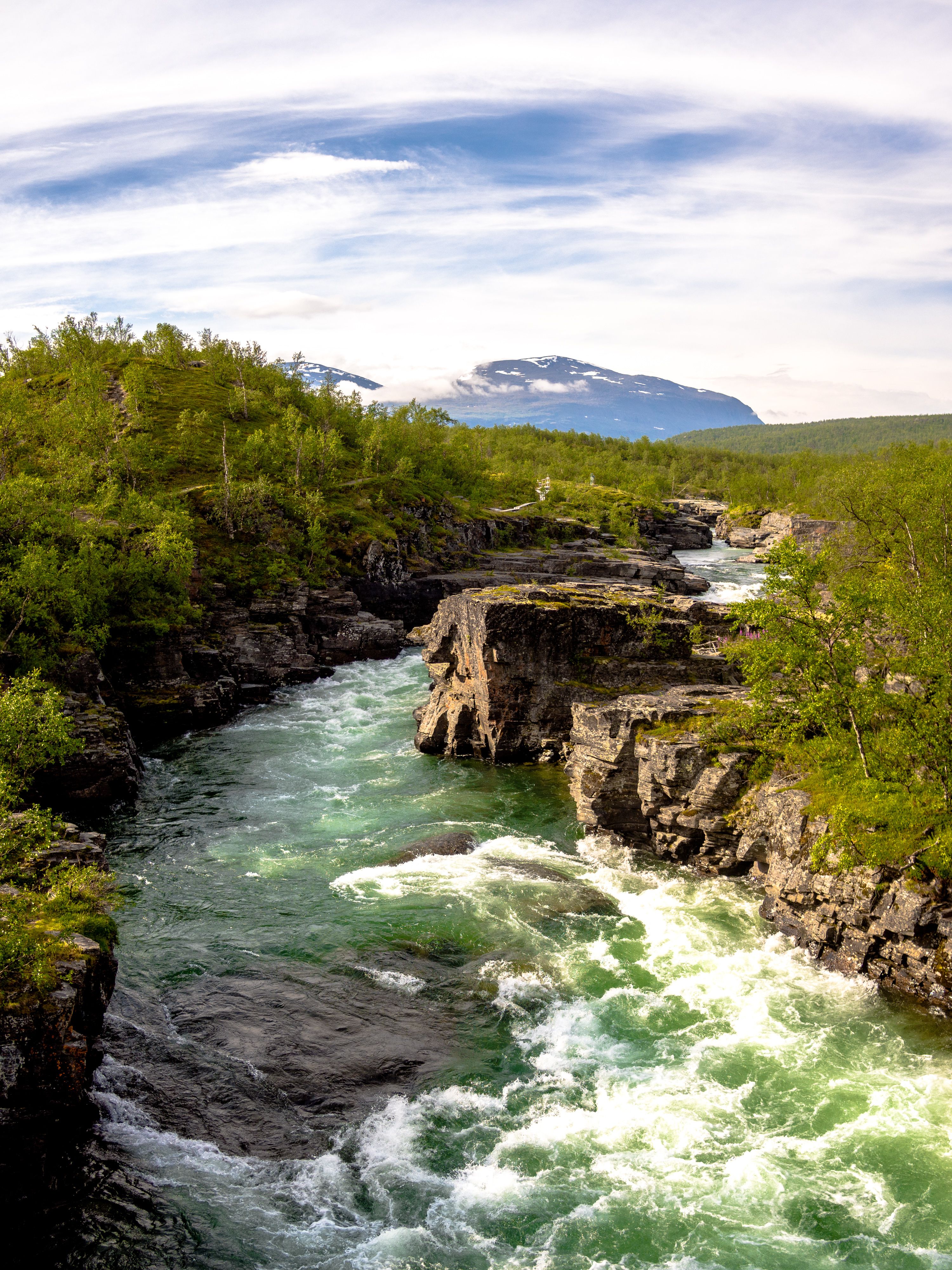 En fors med grönt klart vatten med klippor på båda sidor. Forsen ringlar sig genom landskapet bort mot ett kalfjäll som syns långt bort