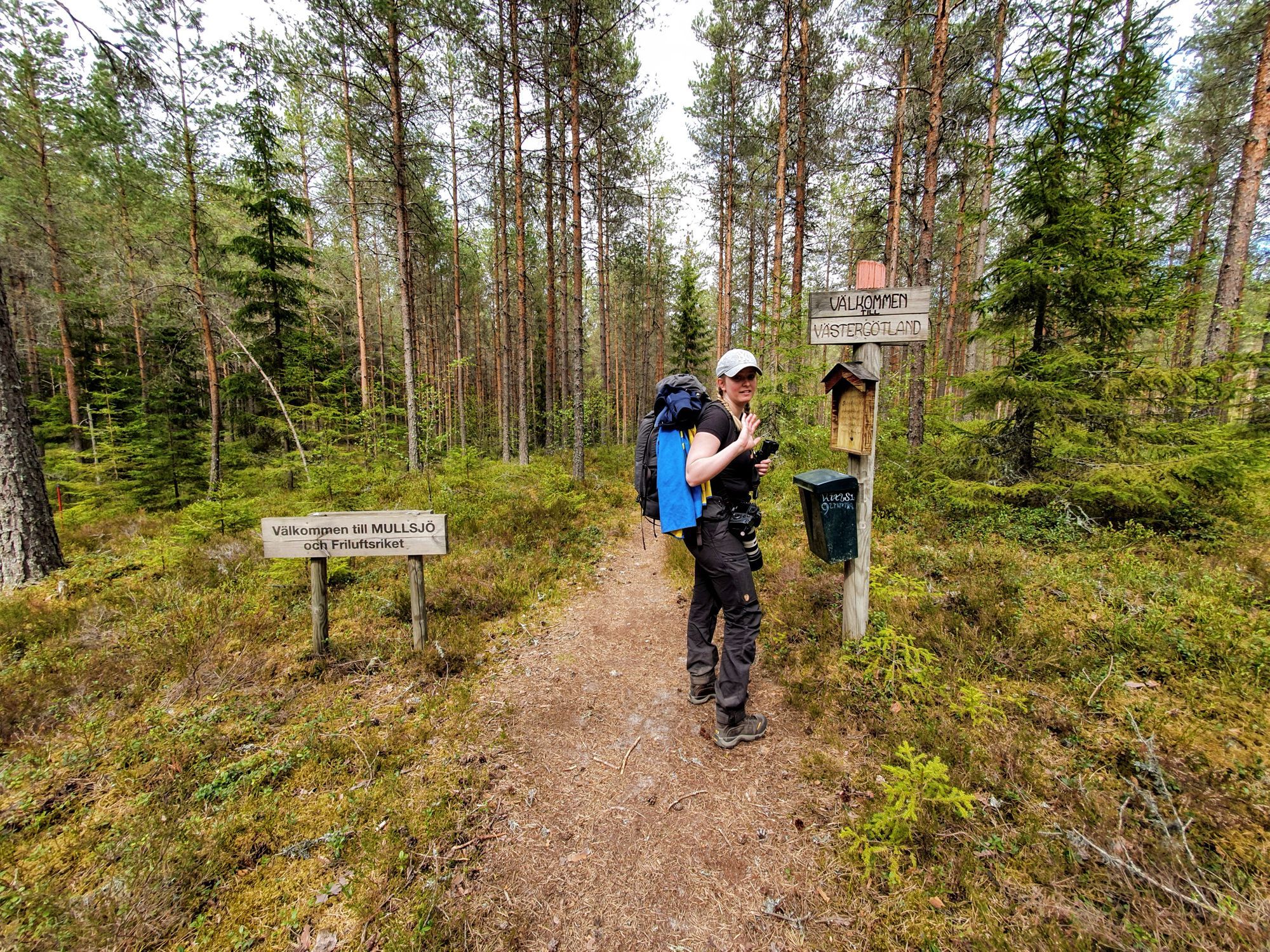 Josefine står på en stig med två skyltar bredvid. En skylt står det “Välkommen till MULLSJÖ och Friluftsriket” på, den andra står det “Välkommen till Västergötland” på.