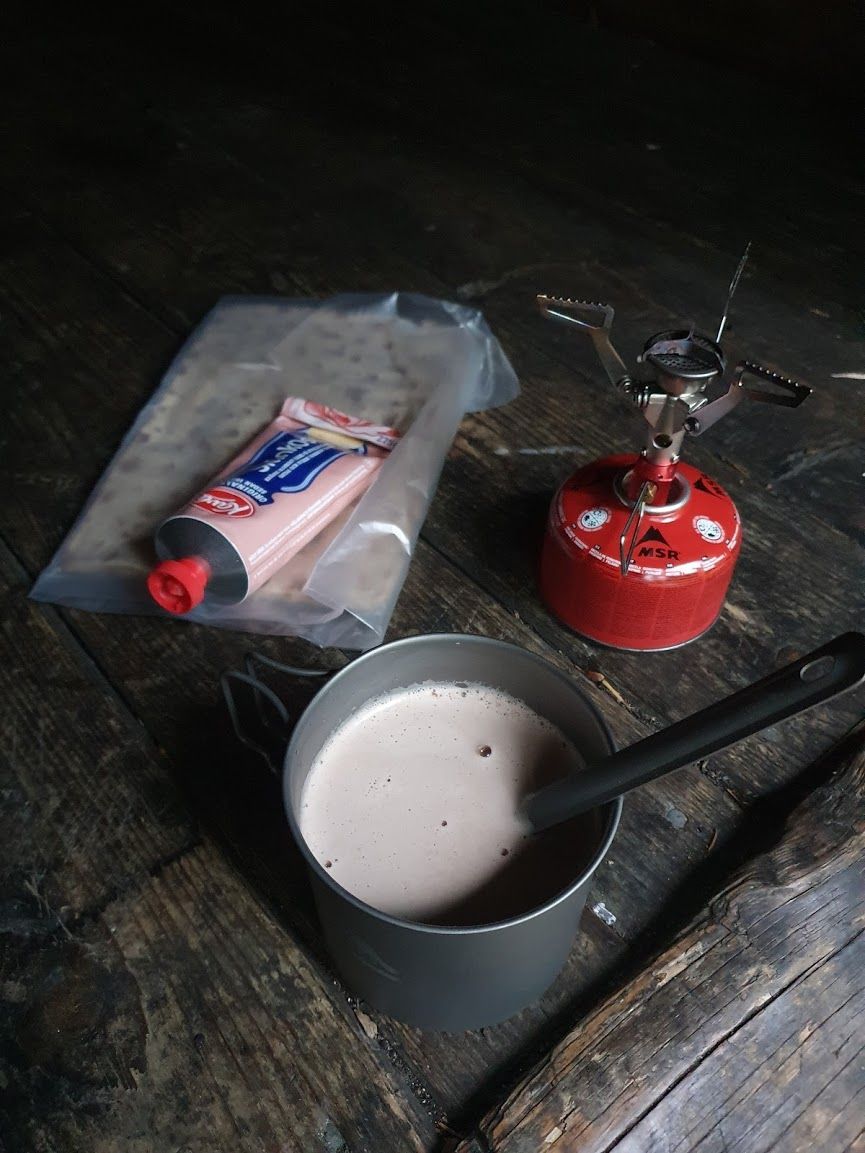 Ett friluftskök med en kastrull fylld med varm choklad, bredvid ligger tunnbröd och mjukost.