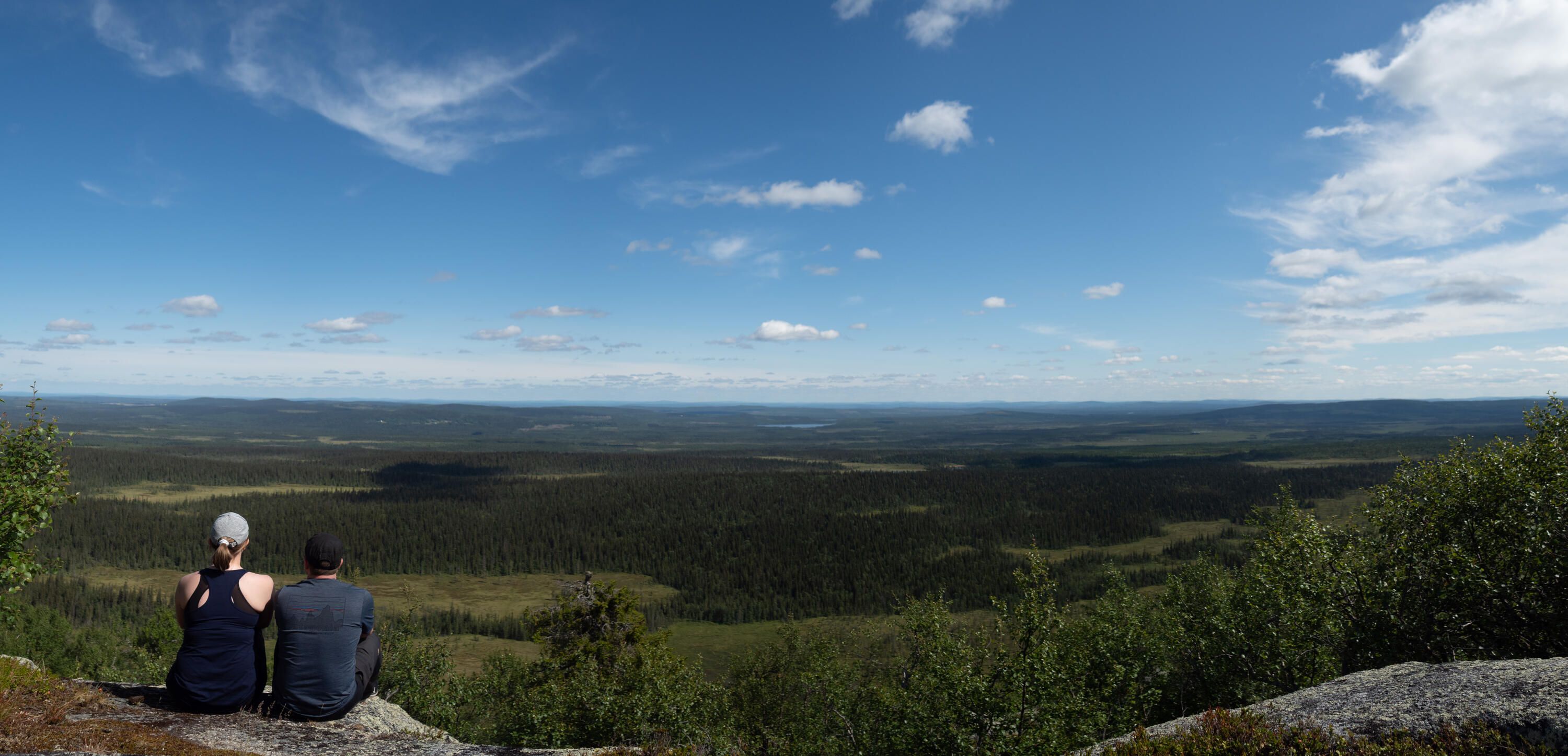 Två personer sitter på en klippa och tittar ut över ett skogs- och myrlandskap