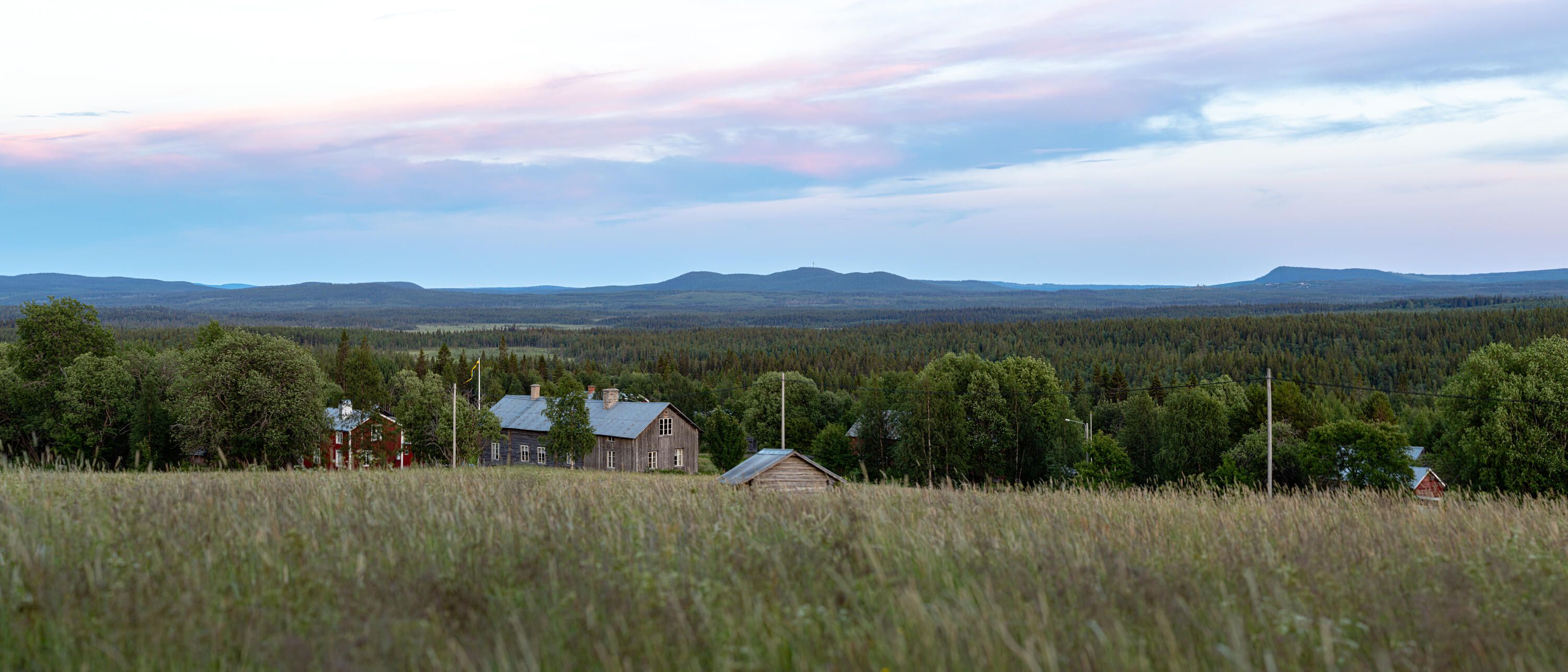 Utsikt över ett landskap belyst av kvällssolen som färgar molnen och himlen i magenta till blått. I mitten på bilden syns några gamla trähus.