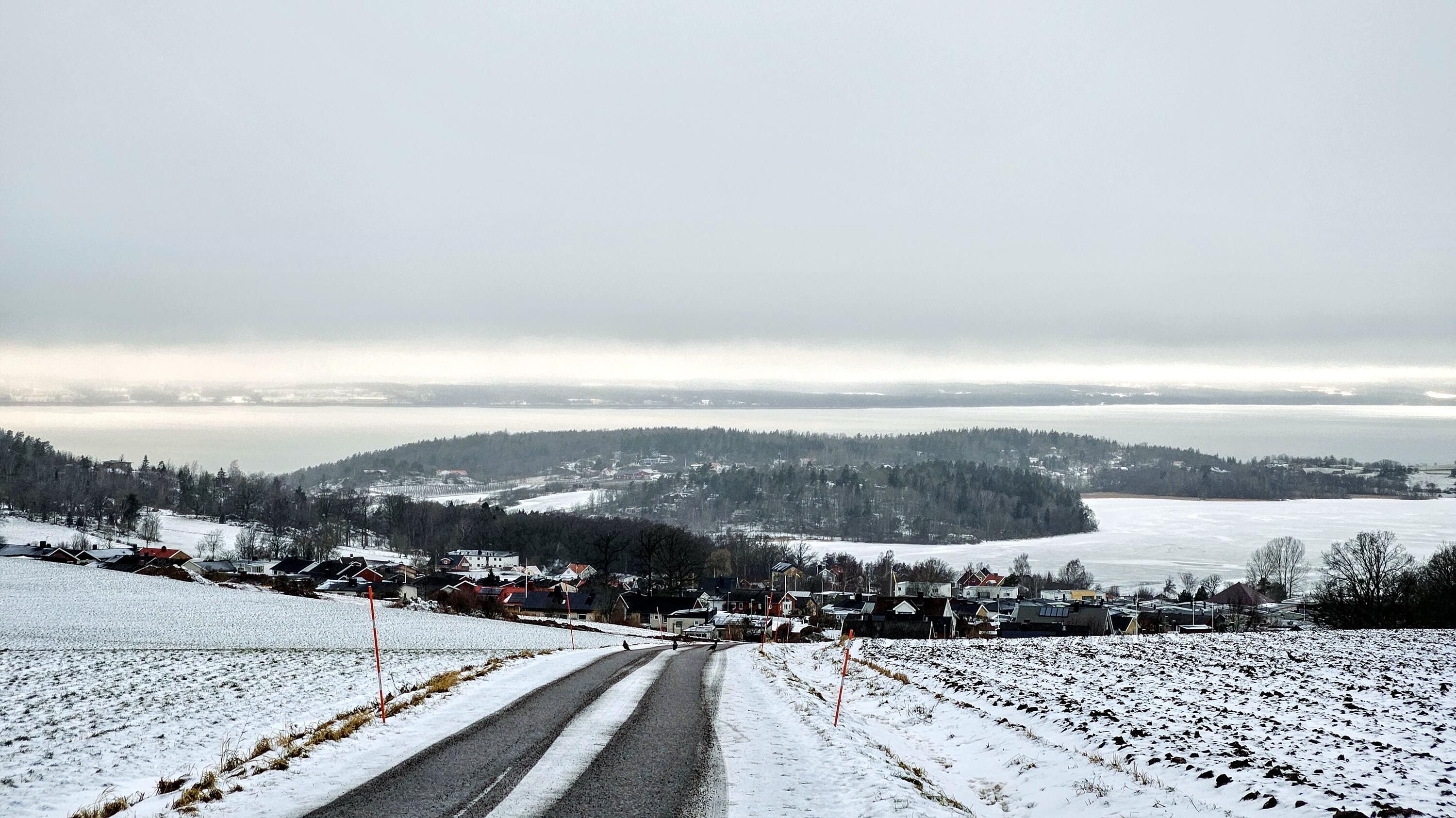 Ett grått vinterlandskap sett från toppen av Gestra ovanför Kaxholmen, i förgrunden syns en grusväg som går nedför berget med åker på båda sidor. Precis nedanför kullen syns hus i Kaxholmen. Bortom det syns Landsjön och Vättern med bara en liten bit land som skiljer dem åt. Landsjön är helt frusen. Molnen hänger lågt