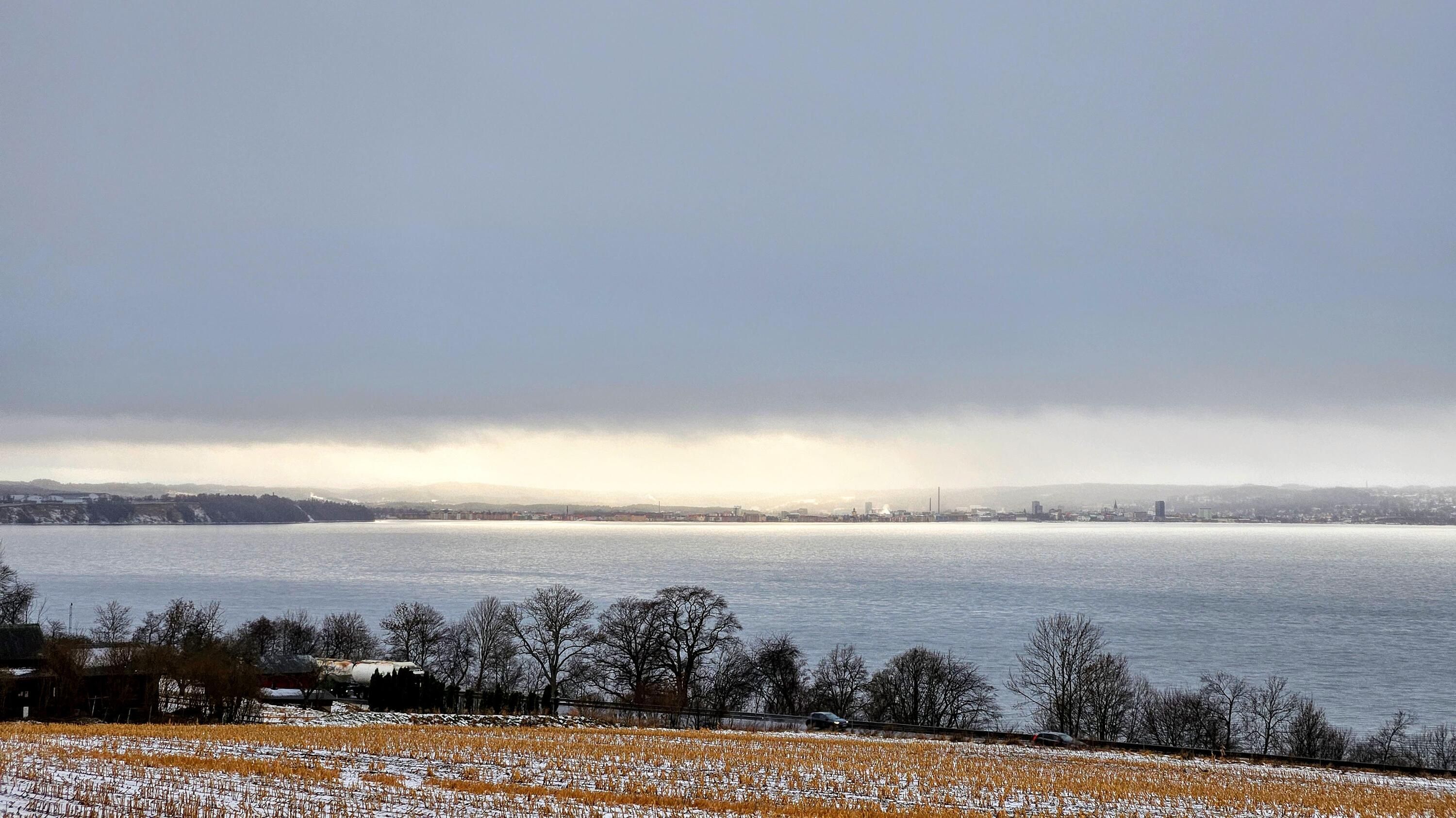 Jönköping sett från andra sidan vattnet. Hela himlen är täckt av ett lågt grått molntäcke utom just vid Jönköping som lyser varmt av solljuset.
