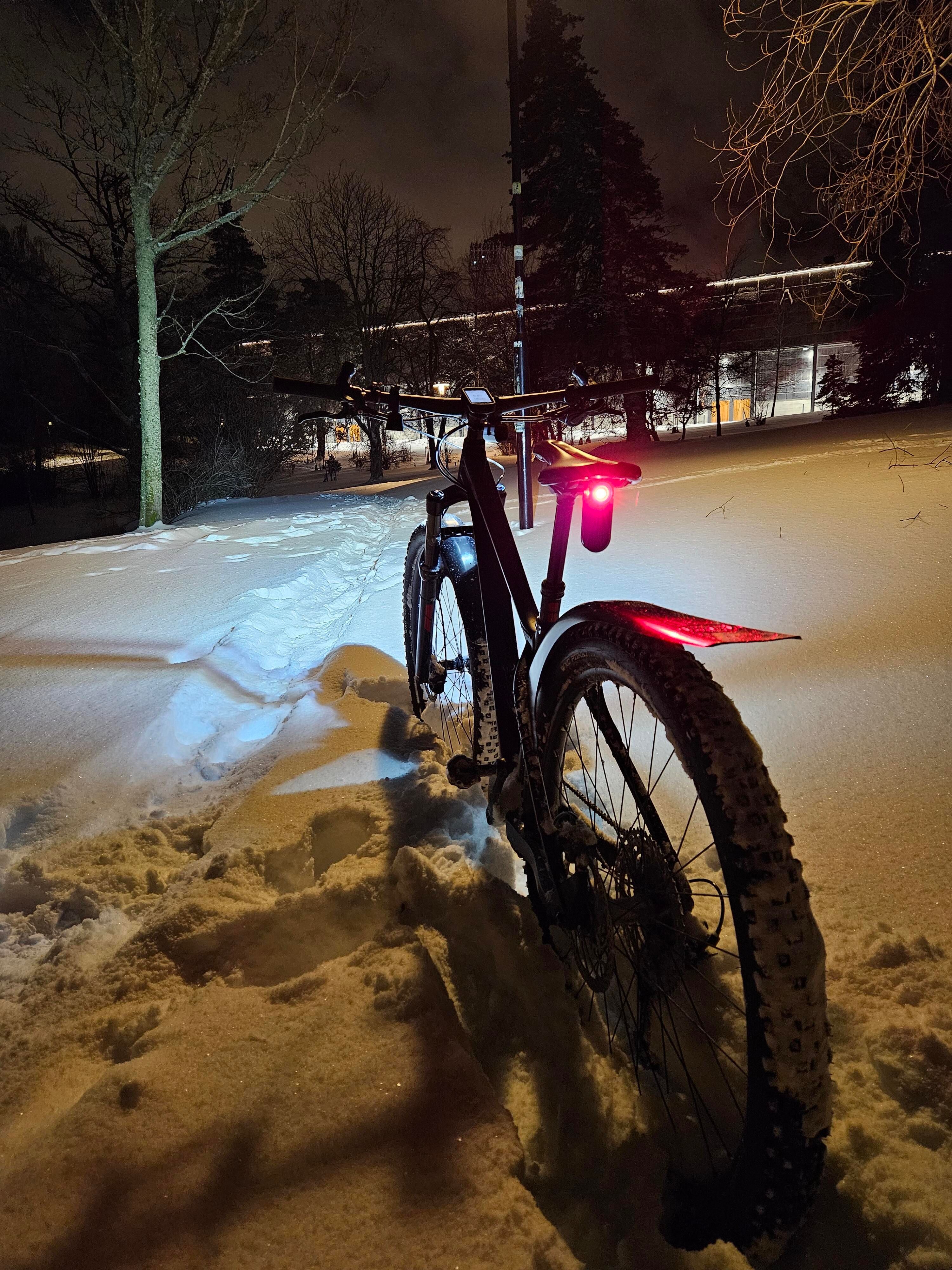 En stigcykel sedd bakifrån som står i djupsnö. Framlampan med starkt vitt sken lyser upp snön och träden framåt.