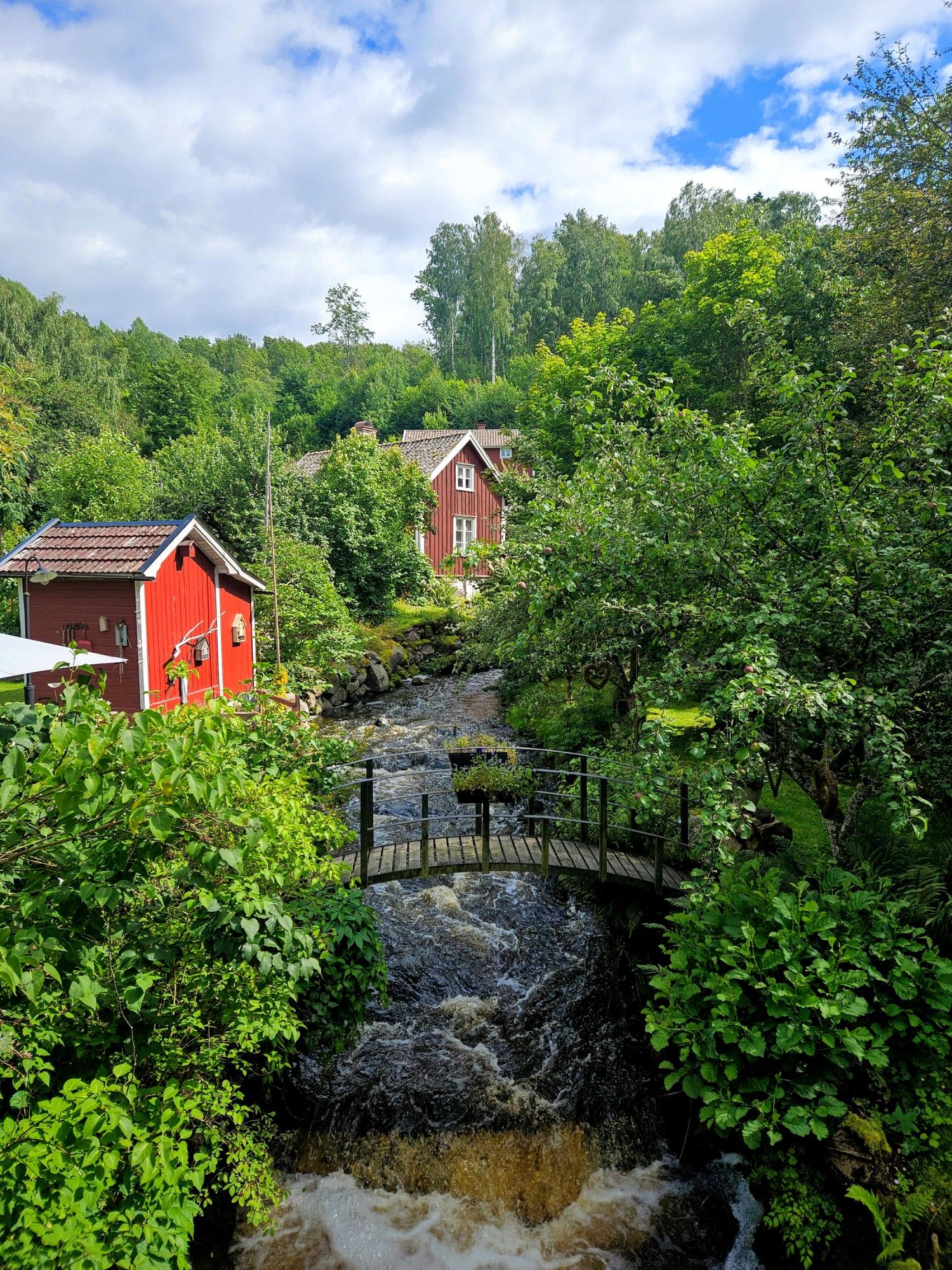 En å som rinner precis intill några faluröda hus. På båda sidor av ån är det mycket buskage och träd. En liten gångbro går över ån.