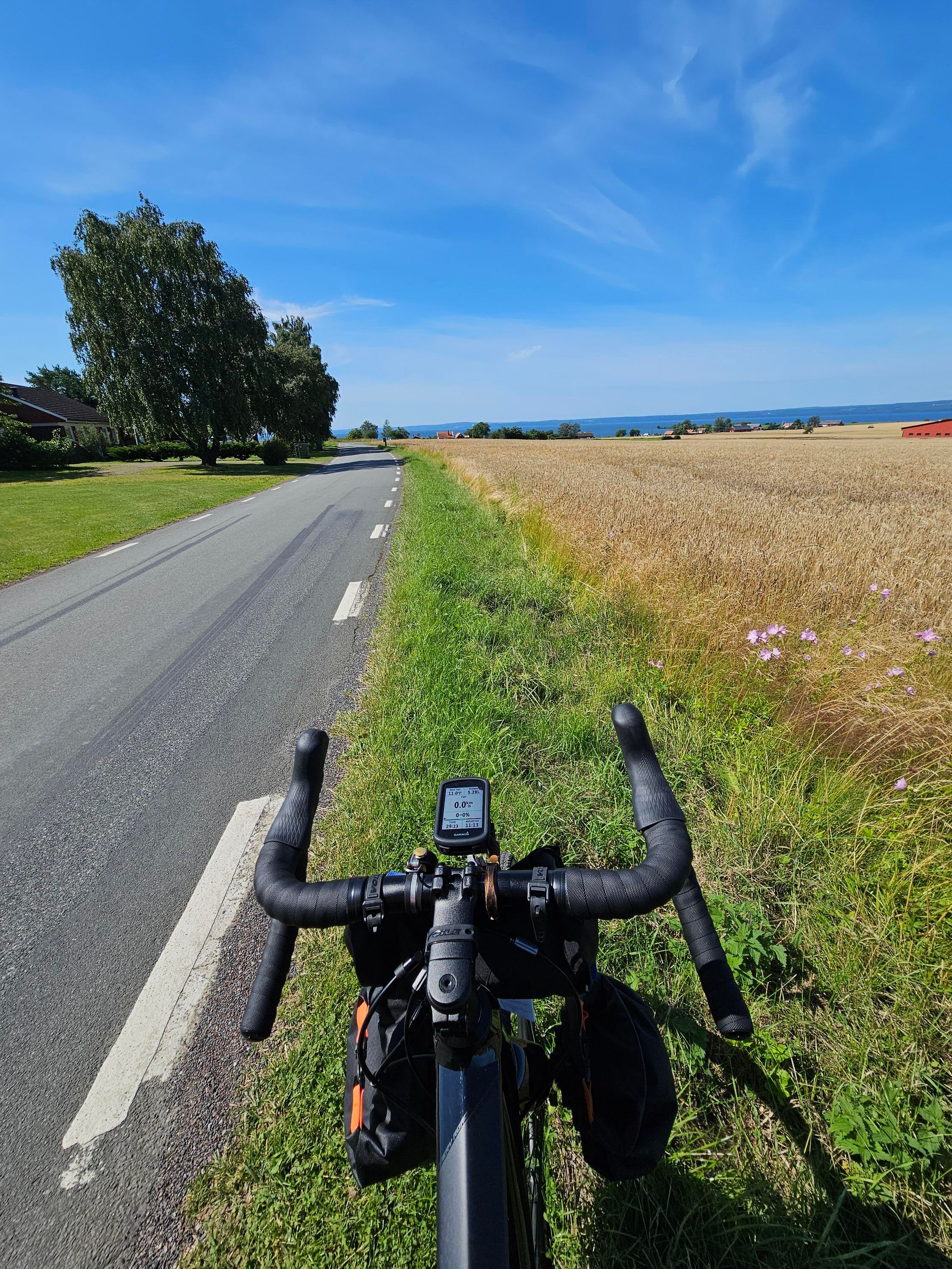 En cykel som står vid sidan om en väg intill en åker. Längre bort skymtas sjön Vättern under en klarblå himmel.
