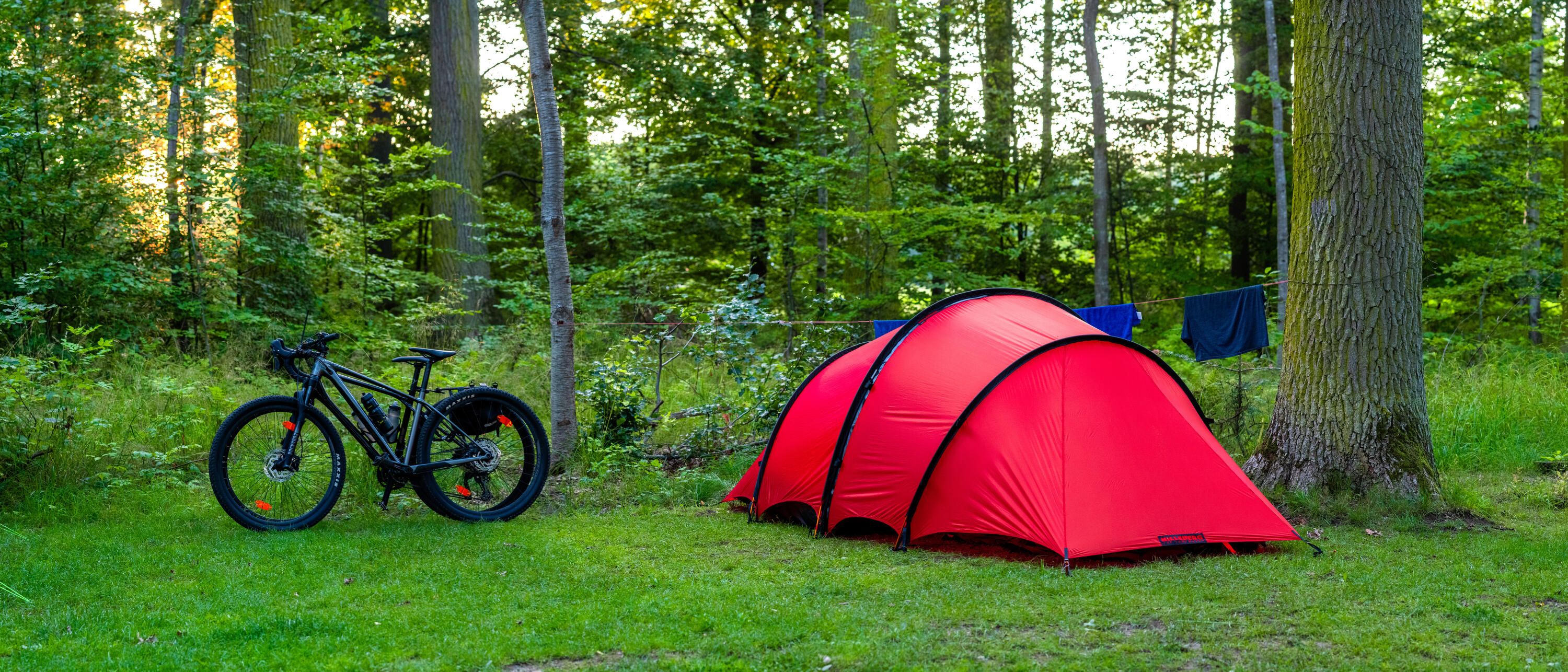 Ett rött tunneltält på en klippt gräsmatta. Intill står två cyklar lutade mot varandra. Bakom tältet skymtas en tvättlina som det hänger några blåa plagg på.