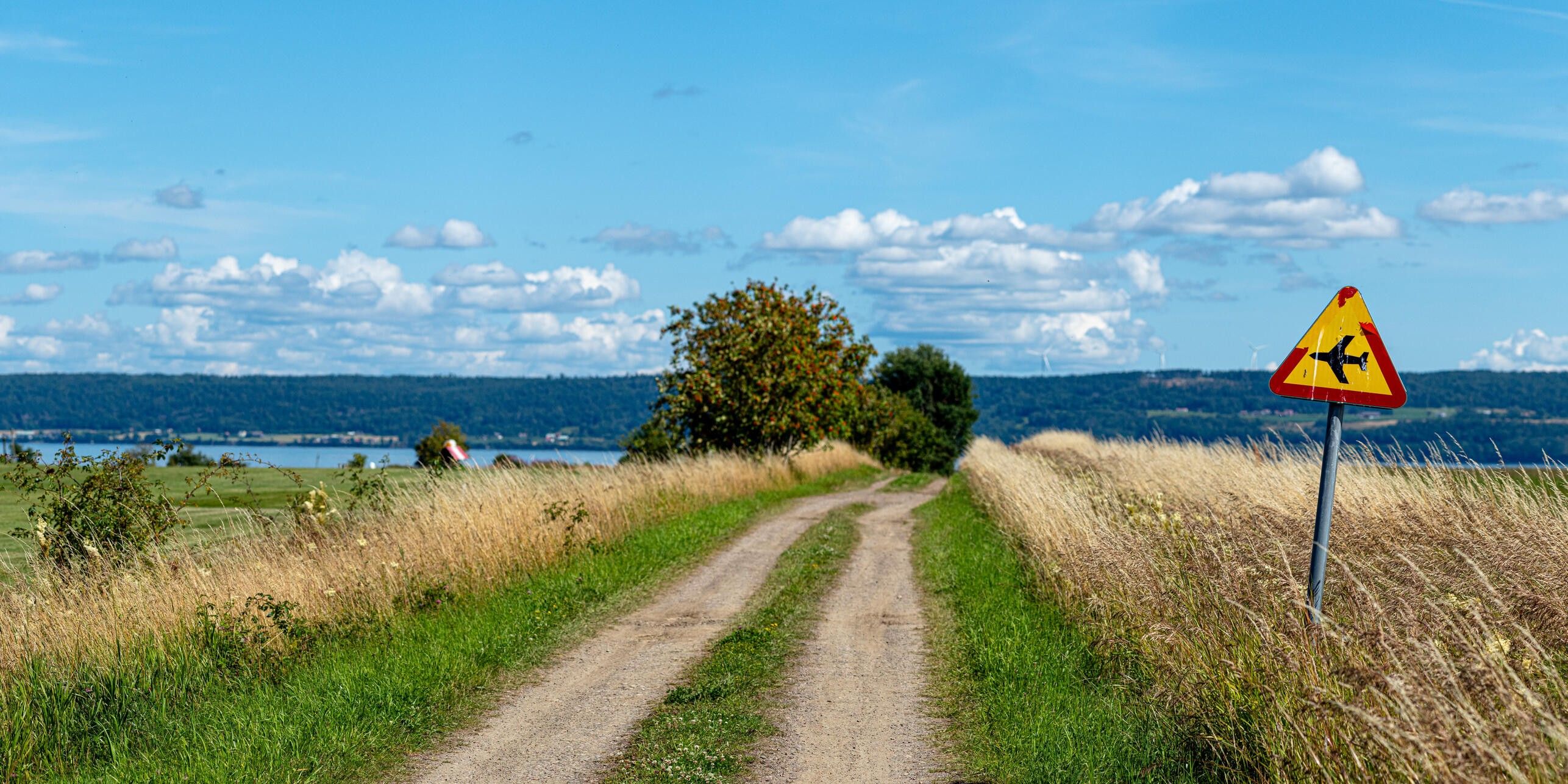 En jordväg med högt gräs på båda sidor som går bort mot ett litet backkrön. Vid sidan om vägen är det en varningsskylt med ett flygplan på