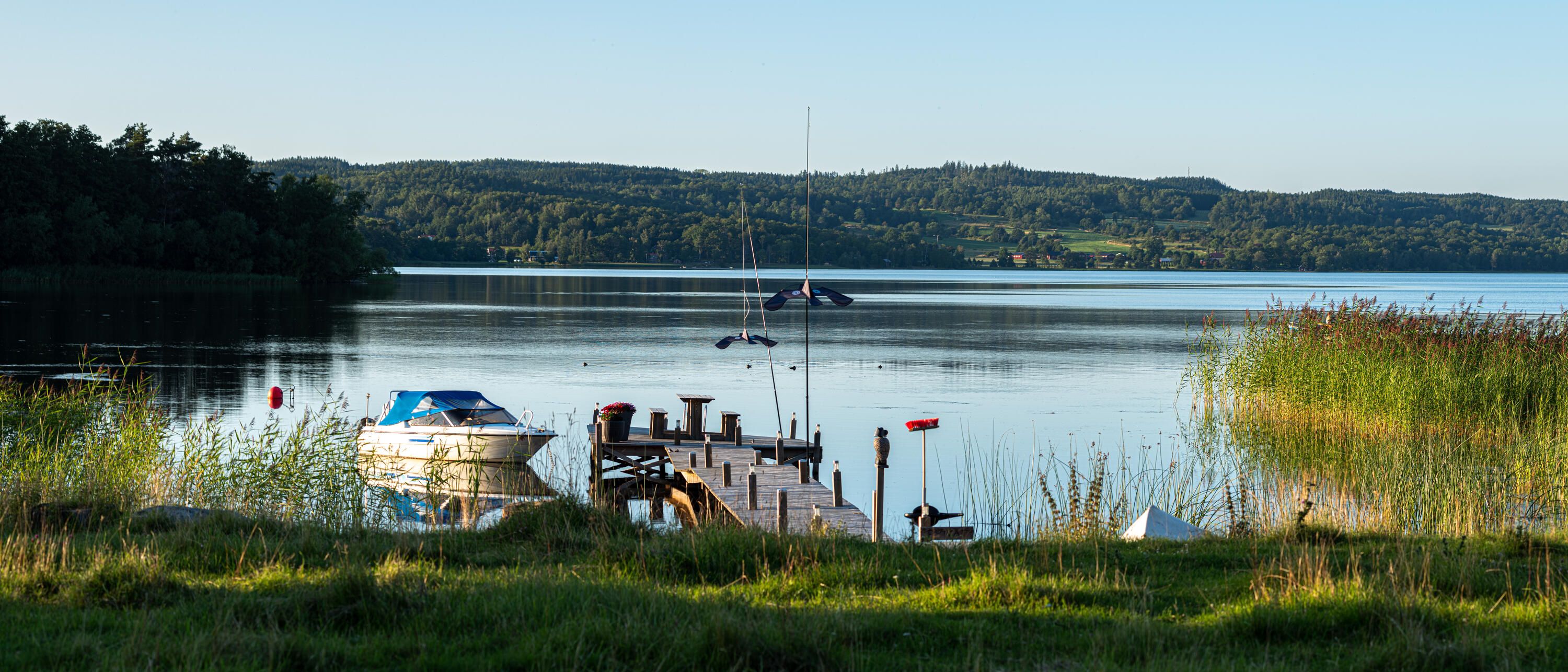 En brygga med en liten fiskebåt i Landsjön. I bakgrunden syns de gröna skogarna på Västa Vätterbranterna.Det sista kvällsljuset träffar bryggan, båten och landskapet och färgar allt gyllene.