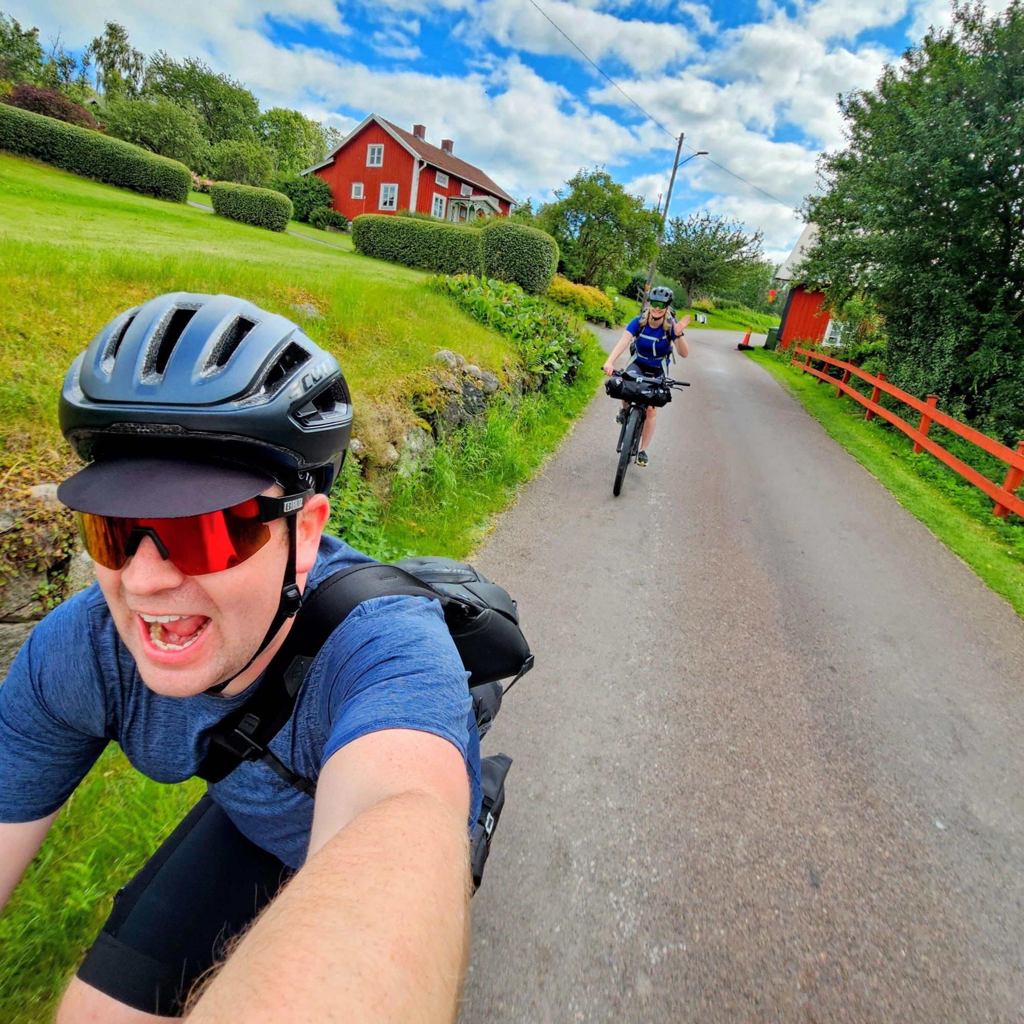 En selfie på Gustav på cykel i hjälm och solglasögon. Gustav har munnen öppen och ser glad ut. I bakgrunden syns Josefine på cykel och vinkar till kameran.