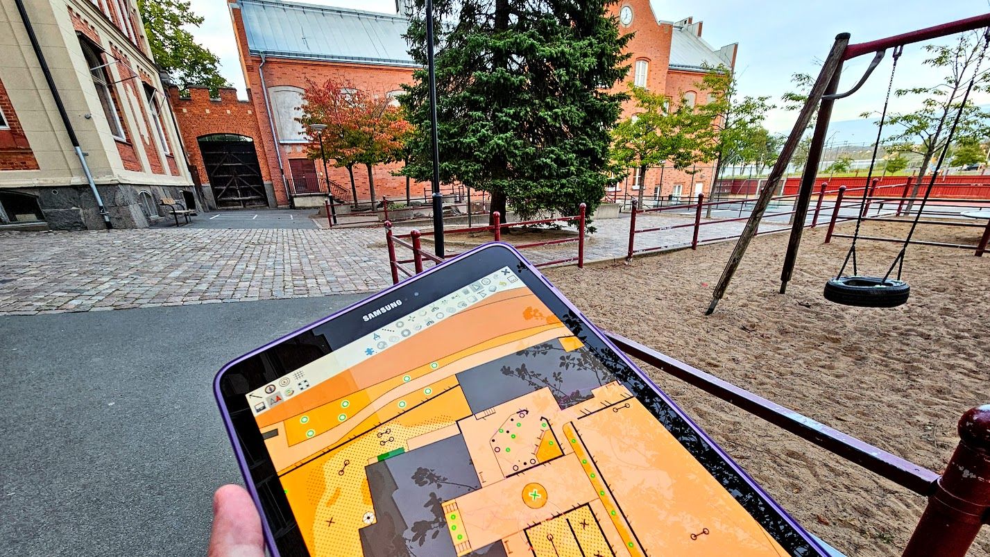 En bild på en surfplatta med en orienteringskarta på och miljön som kartan föreställer i bakgrunden