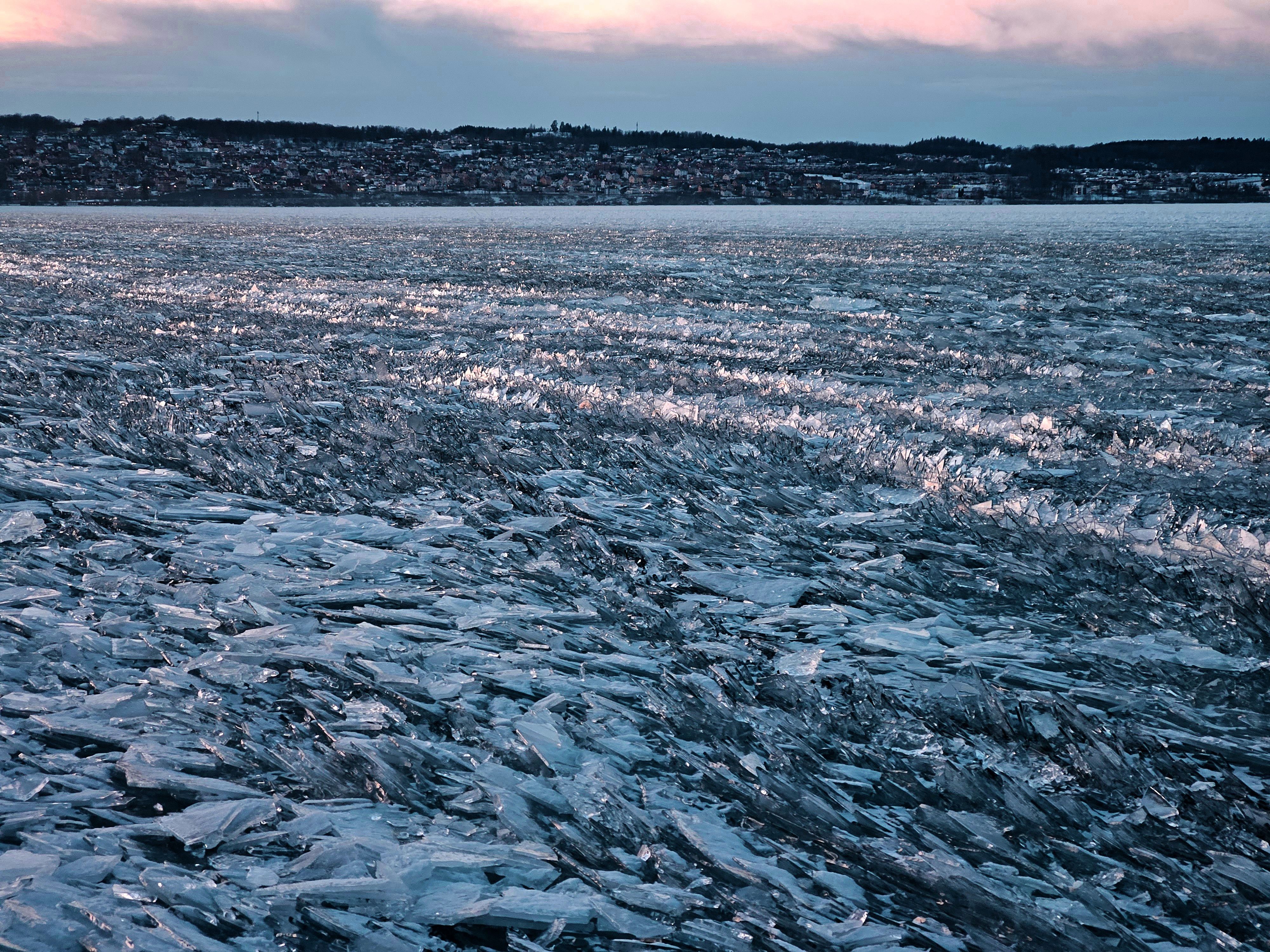 Ett fruset Vättern där isen spruckits och sedan tryckts ihop i flera lager av vassa isflak som sticker åt alla håll. I bakgrunden syns Bymarken.
