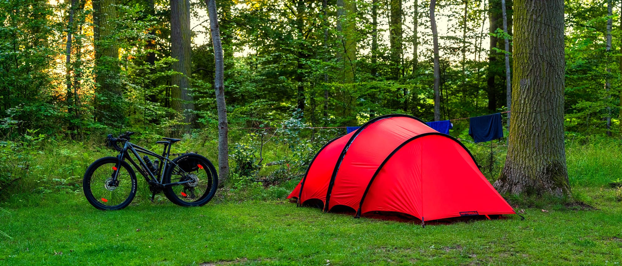 Ett rött tunneltält på en klippt gräsmatta. Intill står två cyklar lutade mot varandra. Bakom tältet skymtas en tvättlina som det hänger några blåa plagg på.