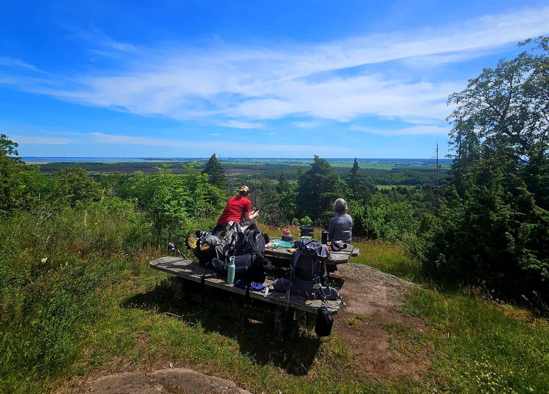 Två personer som sitter vid ett picknickbord med ryggsäckar lutade mot bordet. Det är en utsikt över ett ängslandskap och en sjö i bakgrunden