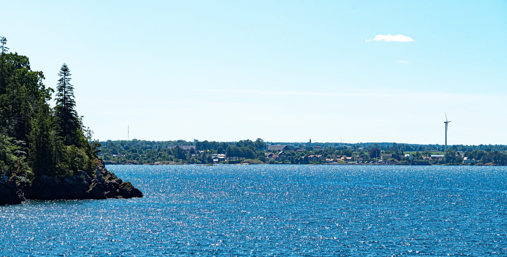 Ett mindre samhälle och en hamn sett på avstånd på andra sidan av en massa vatten. Till vänster i bild syns en klippa med träd på. Vattnet är väldigt blått och reflekterar solljuset i vågorna.
