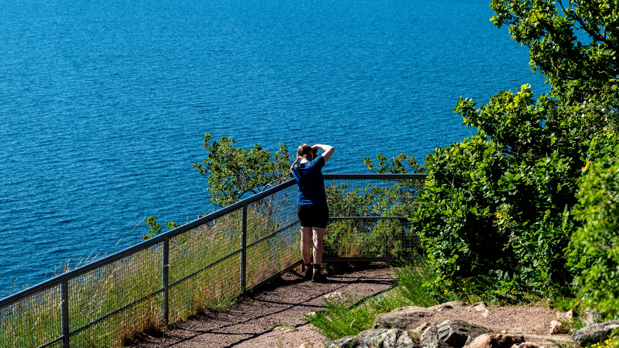 En person som står vid staketet vid en brant och fotograferar ut över vattnet.