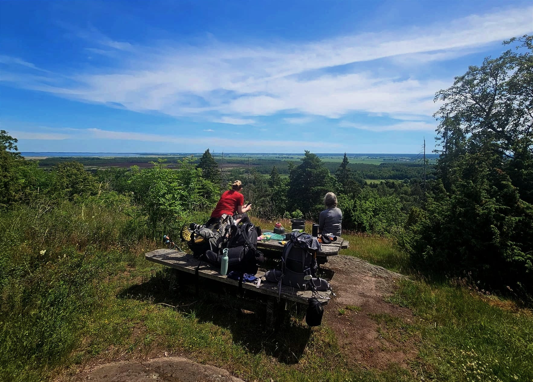 Två personer som sitter vid ett picknickbord med ryggsäckar lutade mot bordet. Det är en utsikt över ett ängslandskap och en sjö i bakgrunden