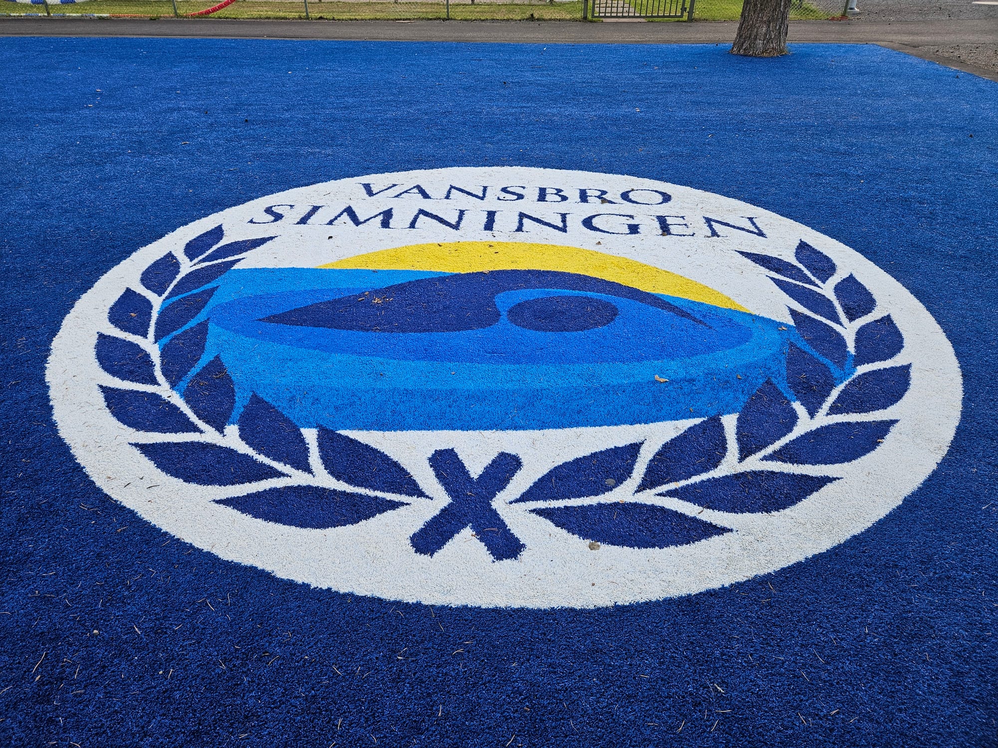 En rund logotyp i marken med texten "Vansbrosimningen" med ett pictogram av en simmare med en krans runt