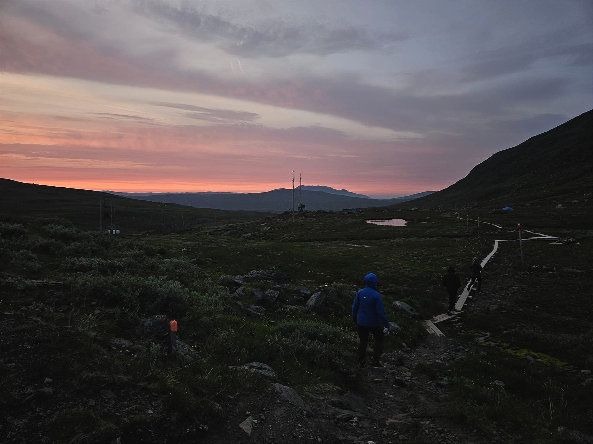 Några personer som går på spångar bort mot en massa tält med det sista av solnedgången i bakgrunden.