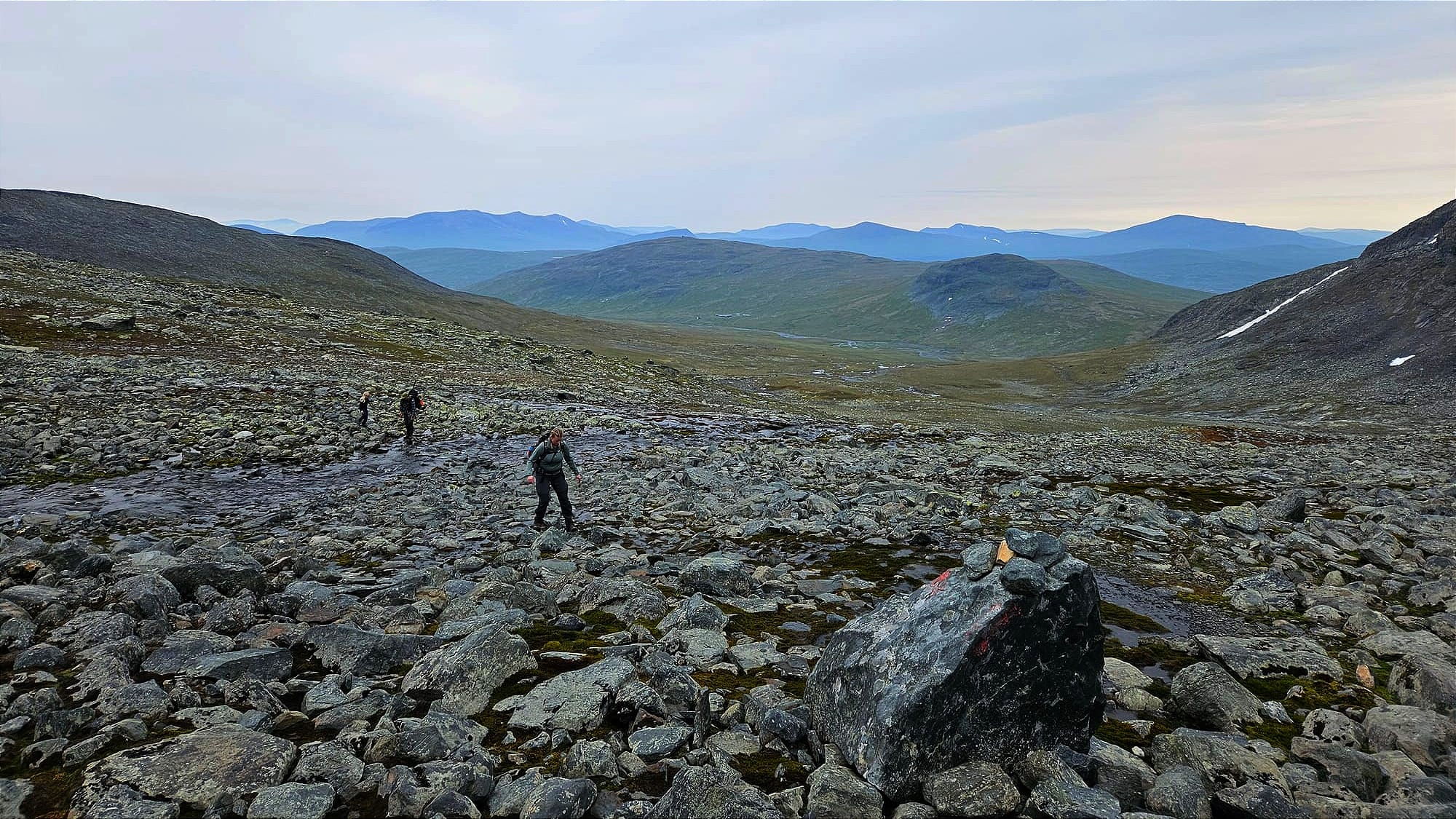 Tre personer som går på en massa klappersten högt upp på ett berg. I förgrunden syns en sten med röda markeringar och i bakgrunden syns en massa berg långt bort.