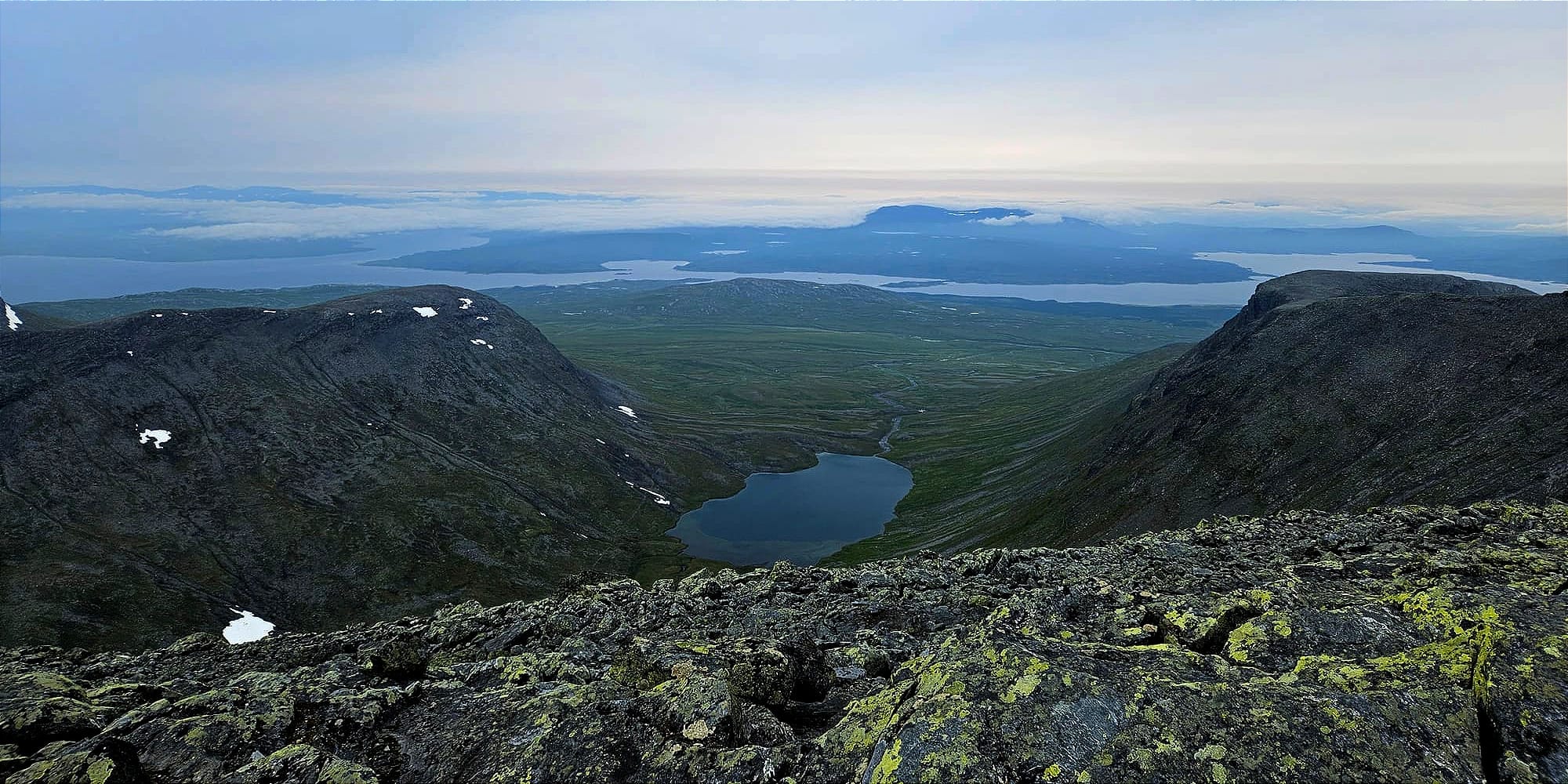 Utsikten från en bergstopp ut över ett platt landskap. Precis nedanför berget syns en sjö. Längre bort är en stor slingrande sjö med låga moln.