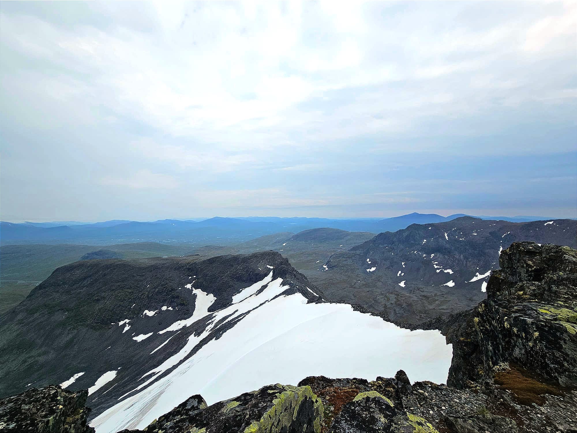 En bergsrygg sedd från toppen av ett berg. Ena sidan av ryggen täcks av en vit glaciär. Långt bort i horsionten syns andra bergskedjor.