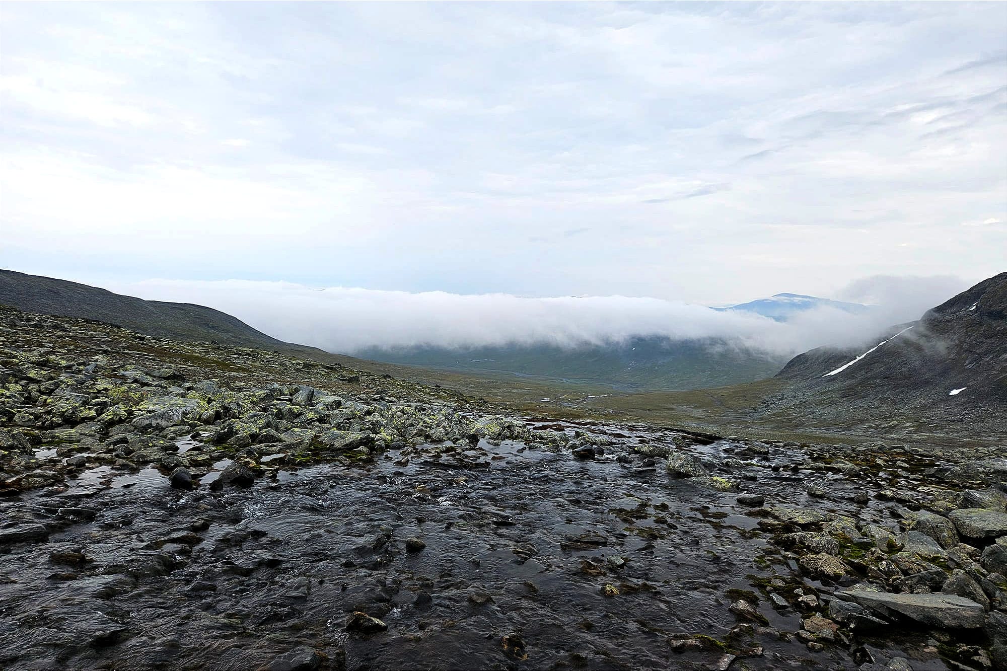 Ett molntäcke som syns över dalen mellan en massa berg. Bilden är tagen mitt eover en stenig bäck som syns i förgrunden