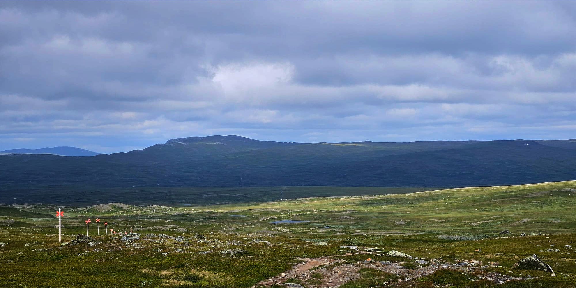 En dal på kalfjället. På andra sidan dalen är ett berg är i skugga och är mörkt blå. I förgrunden badar kalfjället i ljus och lyser i grönt och gult. I gränsen mellan det ljusa och mörka skymtas en liten byggnad.