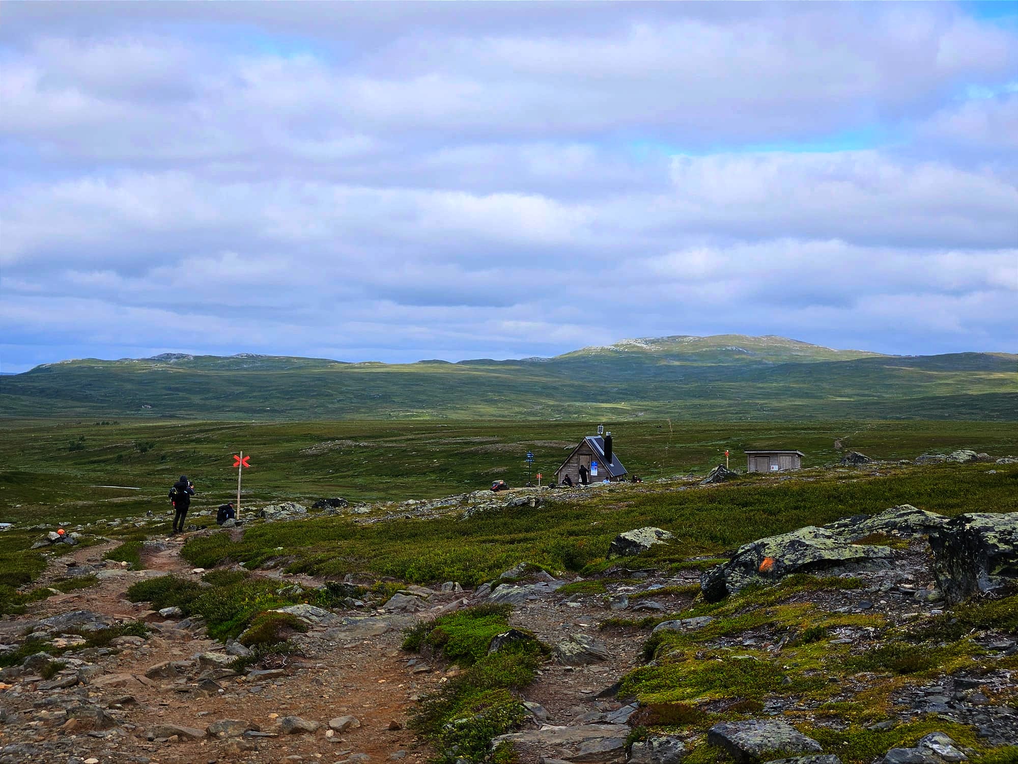 En liten fjällstuga på kalfjället med ett berg i bakgrunden. Solen lyser upp landskapet i fläckar som annars är i skugga.