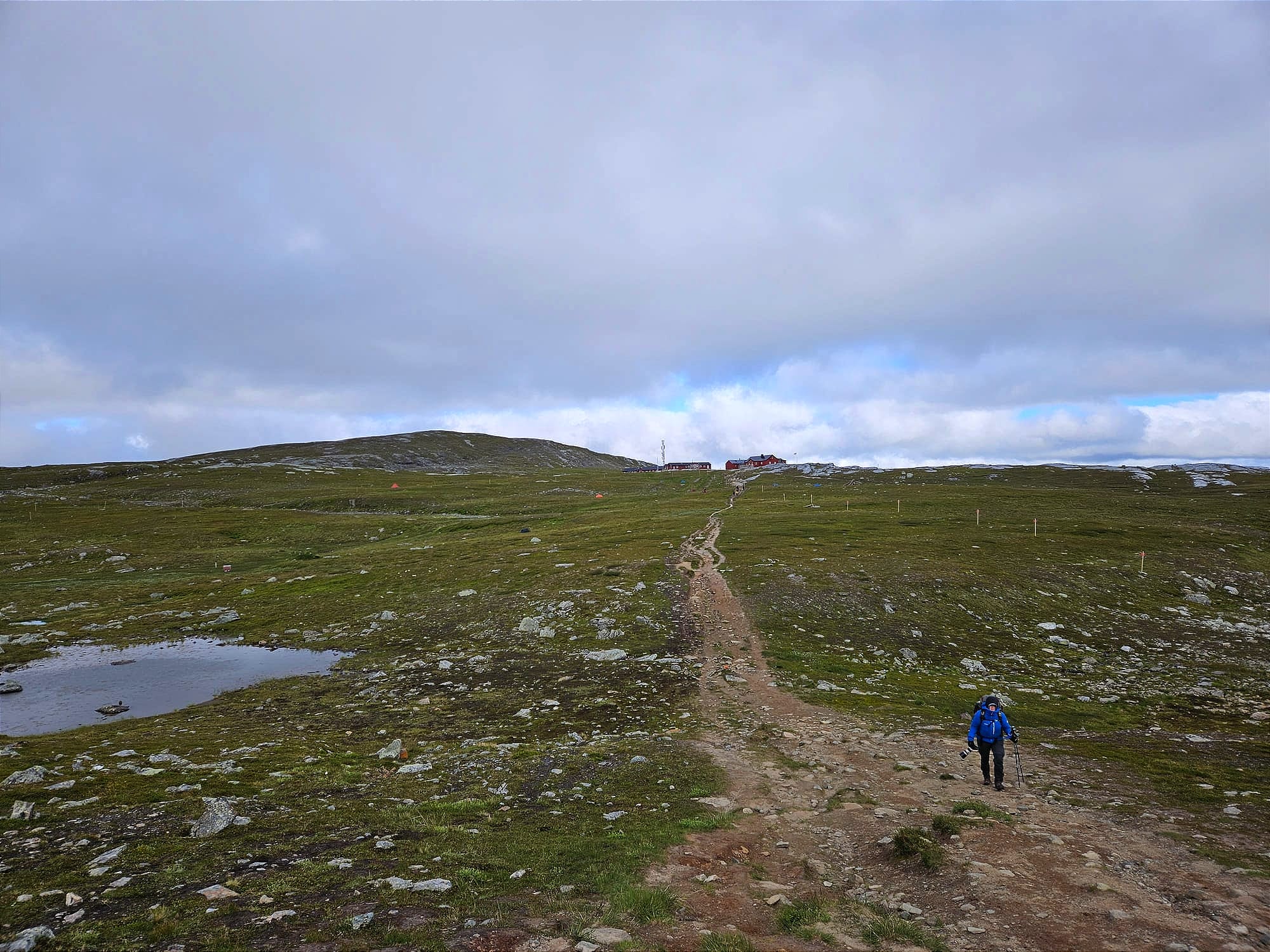 En person går med regnställ och en kamera i handen längs en stig på ett kalfjäll. I bakgrunden i andra änden av stigen syns några röda byggnader och en bergshöjd.