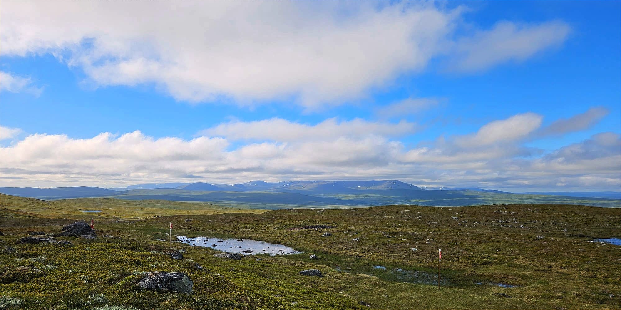 Ett solfläckigt landskap på kalfjället. Marken är grön och långt bort syns en större bergskedja vars toppar är täcka bland molnen.