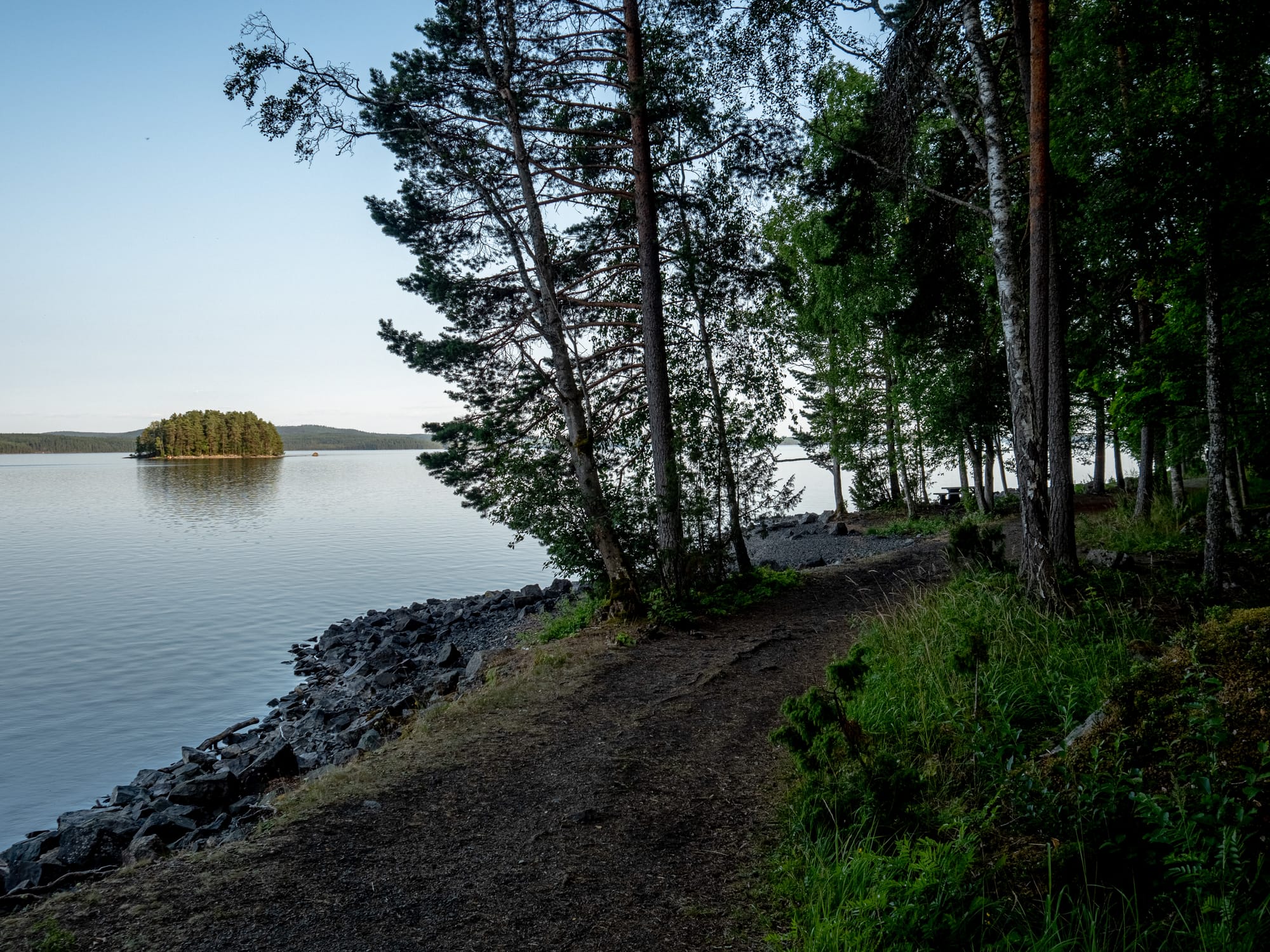 En stenig strand vid en sjö. Intill strandkanten går en stig och det står några träd. Långt ut på sjön syns en skogtäckt ö