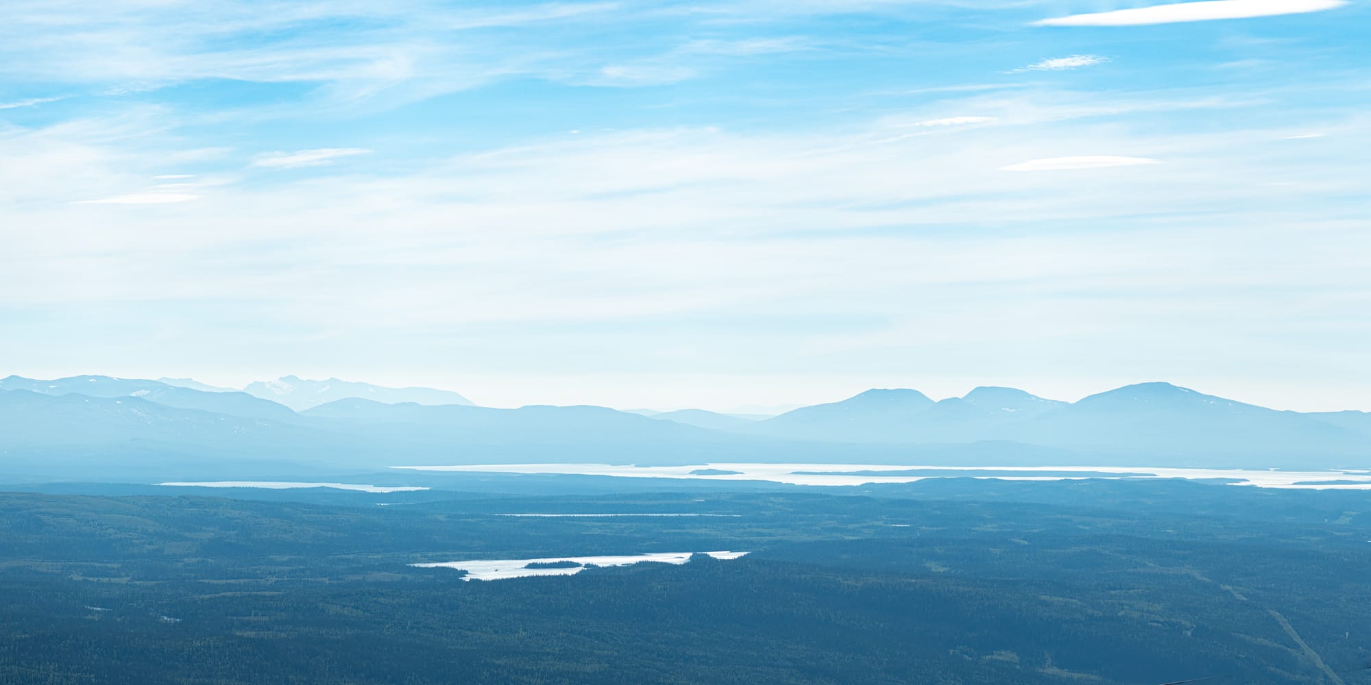 Ett disigt landskap av skogar och sjöar och längre bort flera bergskedjor som syns som siluetter mot den halvmolniga himlen