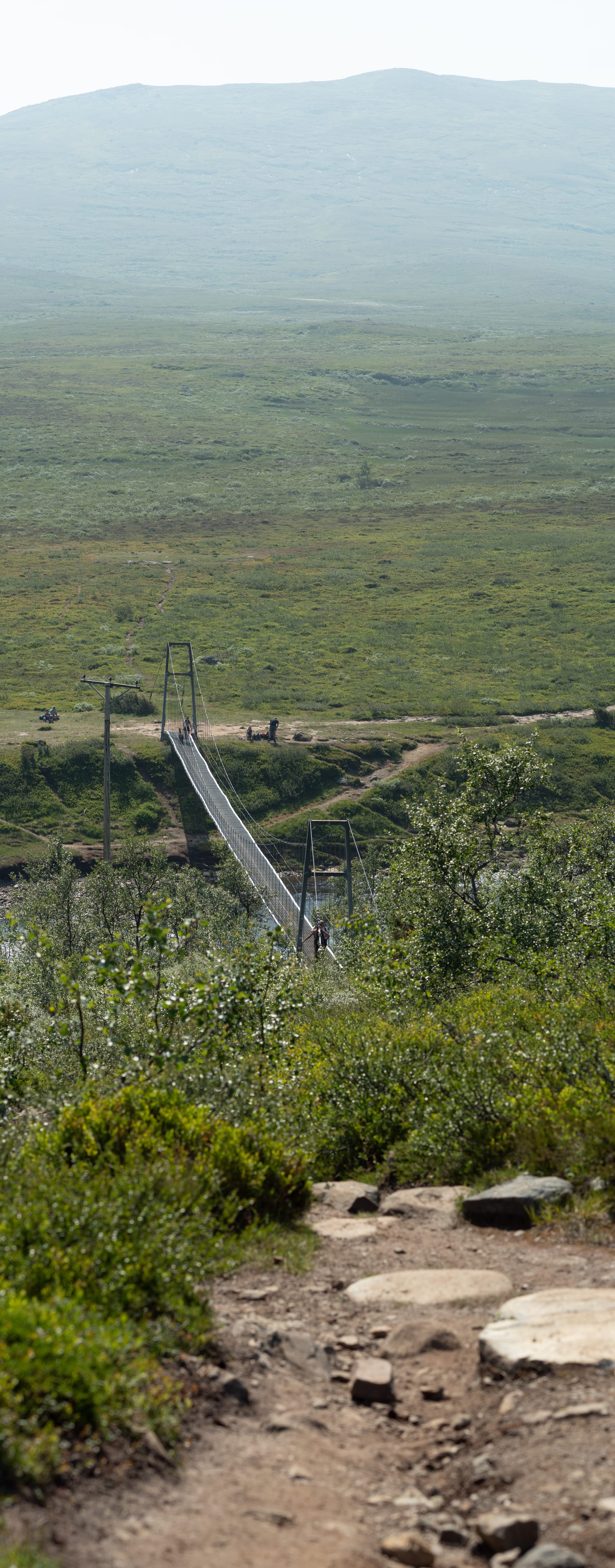 En bro nedanför en kulle med ett kalfjäll i bakgrunden. Förgrunden visar en stig som leder ned mot bron och är helt ur fokus.