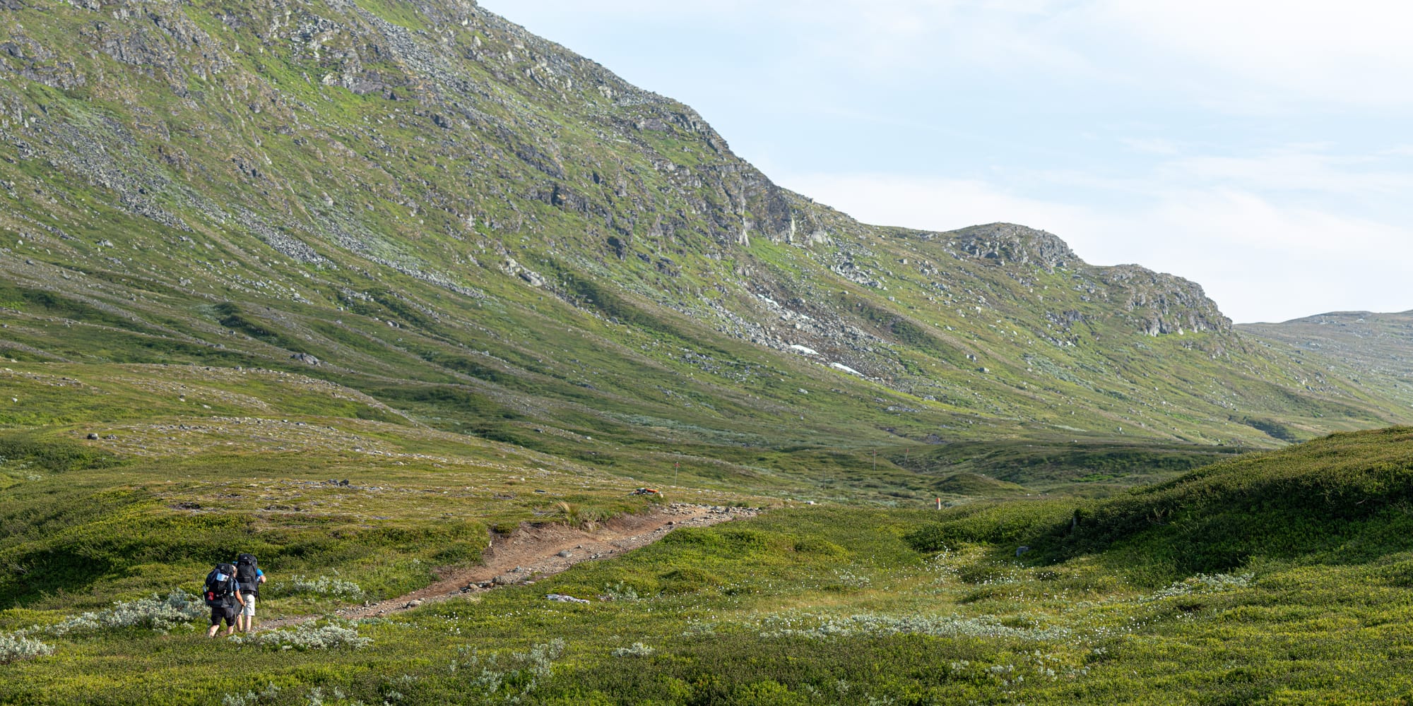 Två personer som går uppför en brant stig. I bakgrunden syns berg som tornar sig över dem och får dem att se väldigt små ut.