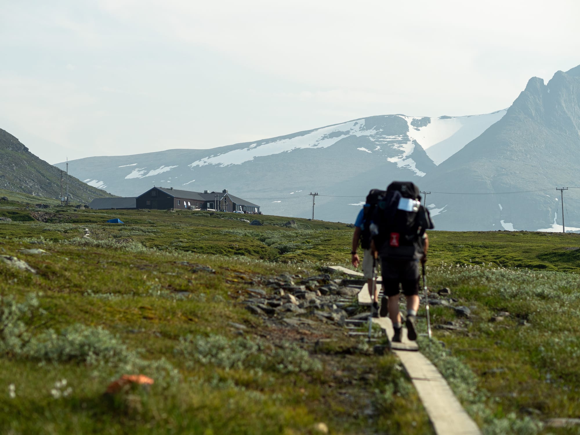 Två personer som går på spångar mot en stor byggnad och en bergskedja i bakgrunden. Fokus i bilden ligger på bakgrunden och personerna är ur fokus.