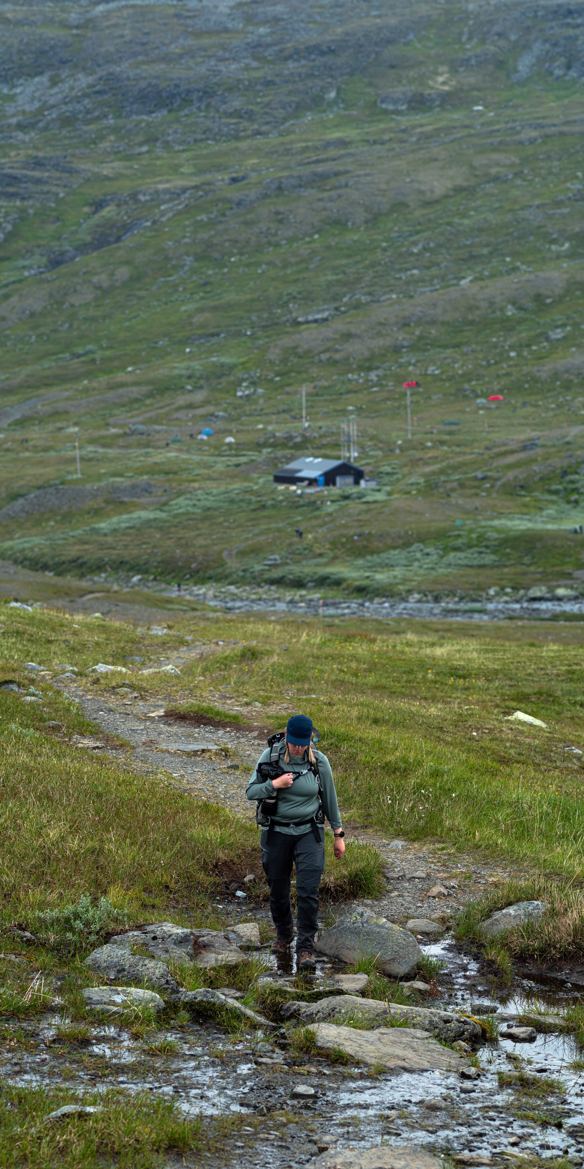 En person som går ner i en stenig bäck med en stig som leder nedför ett berg mot en byggnad som syns långt bak ur fokus