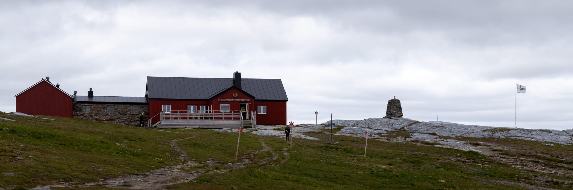 En röd byggnad på toppen av ett stenigt berg. Till höger om byggnaden syns ett stenröse och en vit, gul, blå korsflagga med STF:s logotyp i mitten