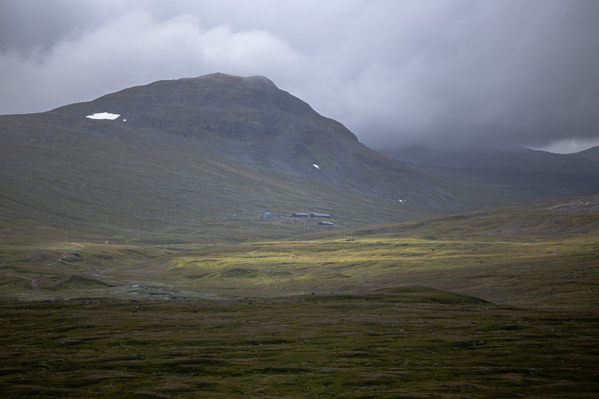 En dal med några byggnader i skugga med några fläckar av solljus. Dalen är fotograferad på ett stort avstånd och höga berg syns i bakgrunden