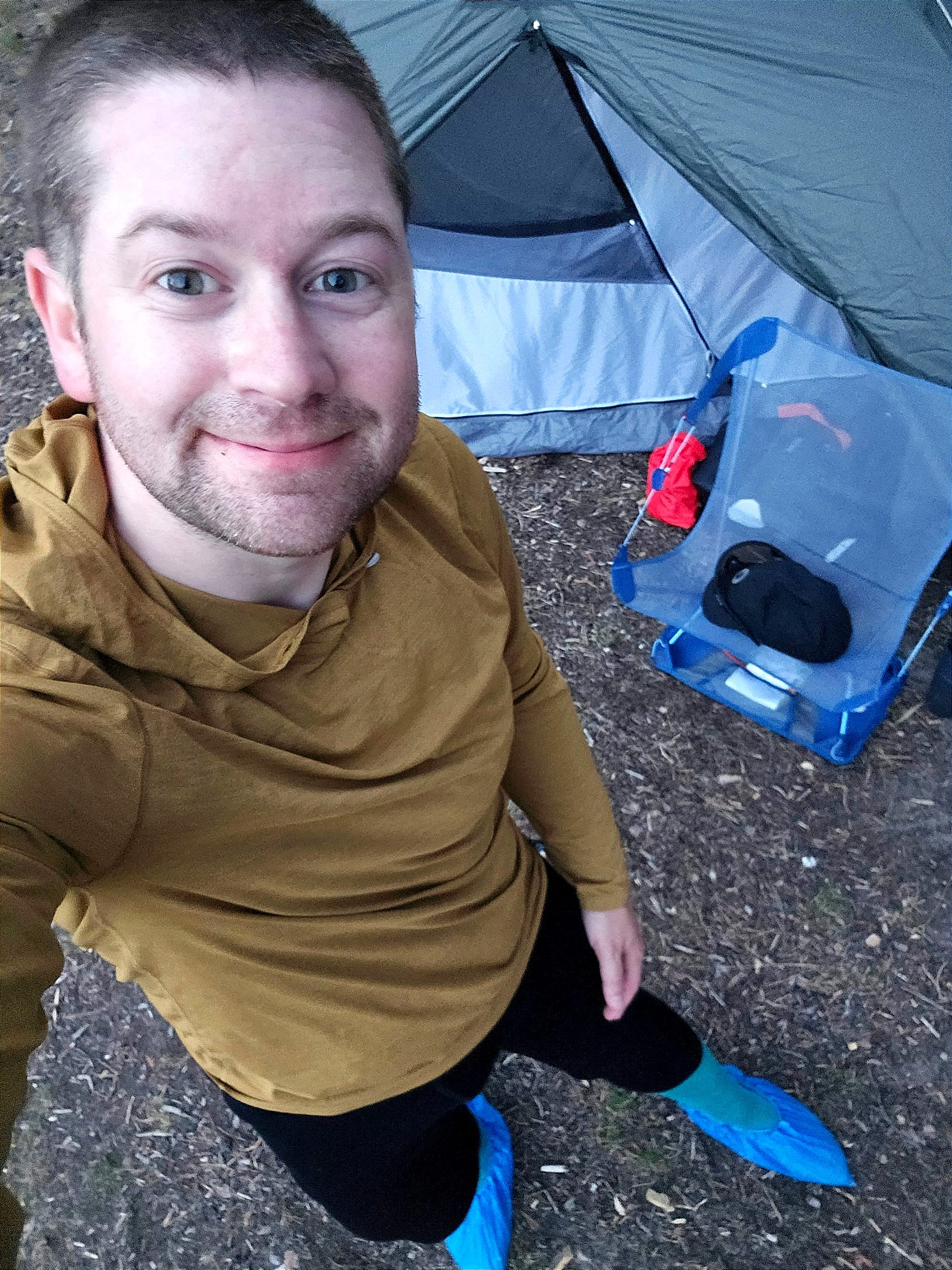 En selfie på en leende person med gul tröja och blå skoöverdrag på fötterna.