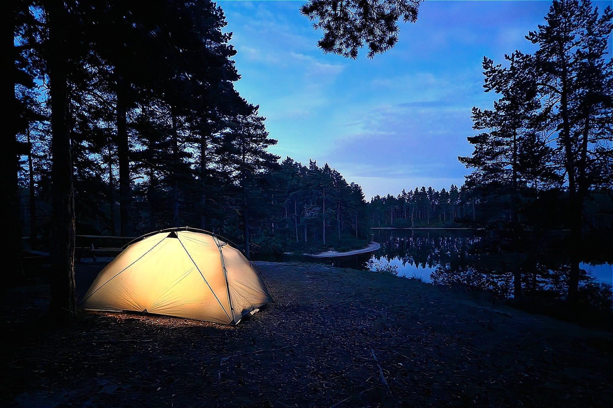 Ett tält på en höjd i en skog med utsikt över en sjö. Tältet är upplyst av en gul lampa inifrån. Skogen och sjön är mörk.