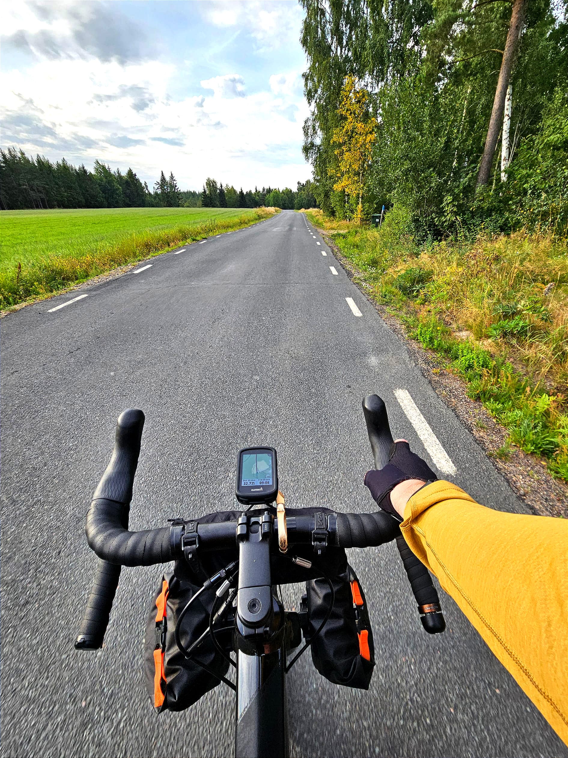 En cykel med bockstyre på en asfalterad väg. En arm med en gul tröja håller i styret. På ena sidan vägen är det en åker och på andra sidan är det skog.
