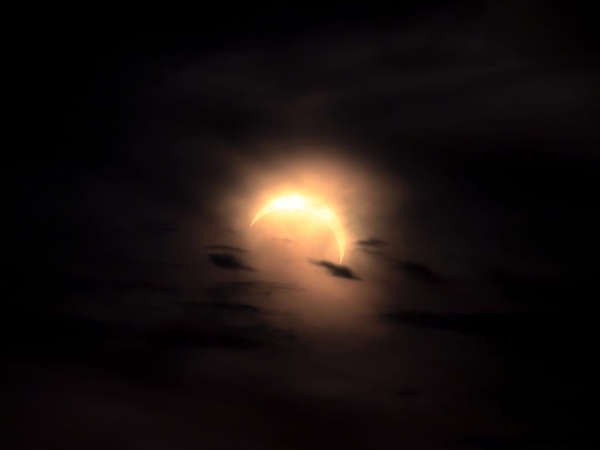 En bild på en solförmörkelse. Månen täcker runt 80% av solen. Bilden är exponerad så att solen är nästan helt vit med ett gult sken runt, resten av bilden är väldigt mörk. Himlen är mörkt grå med nästan svarta moln utspridda.