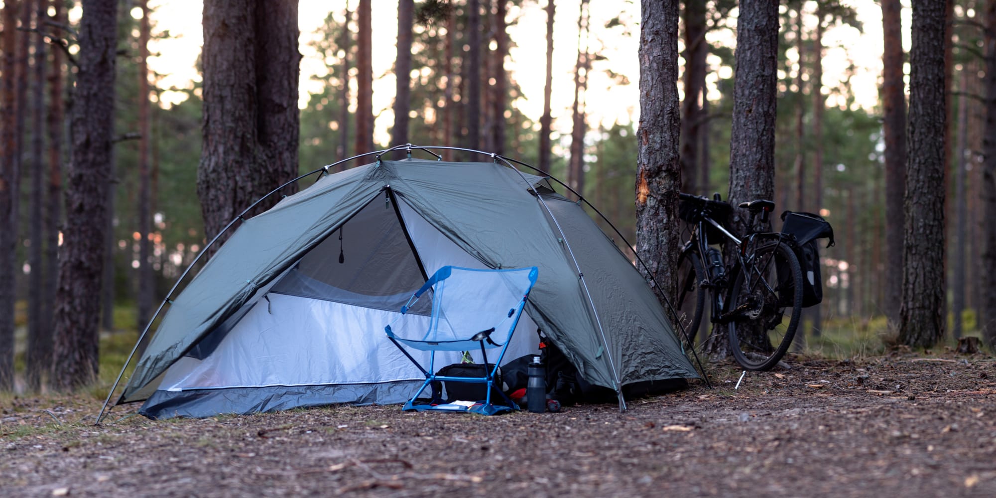 Ett grågrönt tält i skogen med yttertältet öppet på en sida. Framför tältet är en campingstol uppställd och en cykel står lutad mot ett träd bakom tältet.