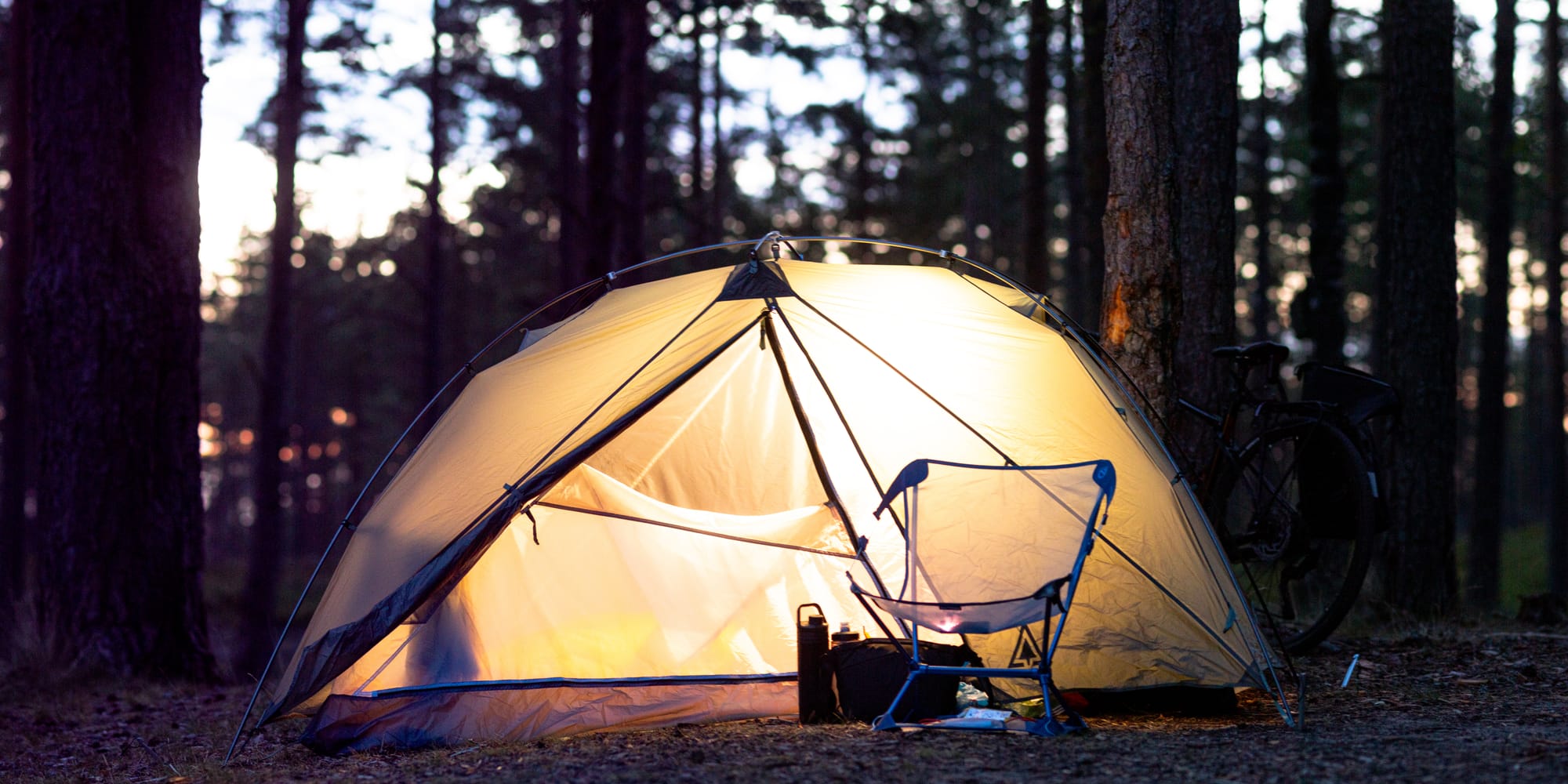Ett tält på kvällen i skogen. Tältet lyses upp gult av en lampa inifrån. En campingstol står framför.