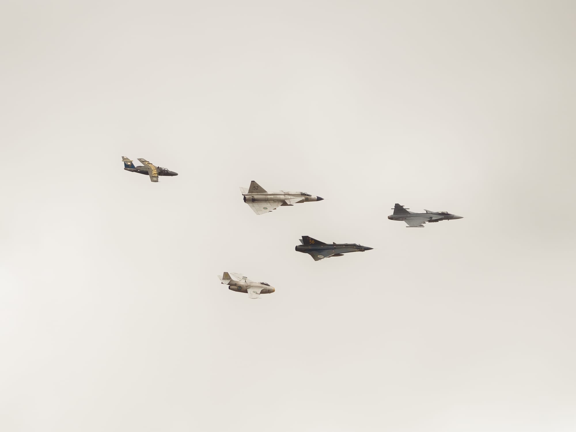 Fem flygplan som flyger i en v-formation. Flygplanen är mestadels stridsflygplan från olika generationer