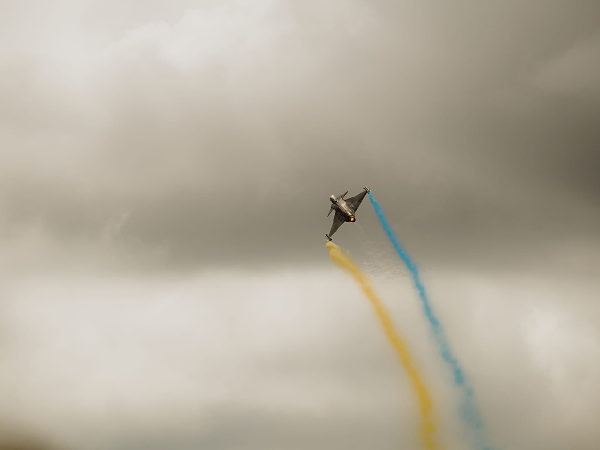 Ett stridsflygplan som släpper ut gul och blå rök. Flygplanet är sett bakifrån och flyger upp mot en grå himmel. Från motorn syns en varm jetström som bryter ljuset